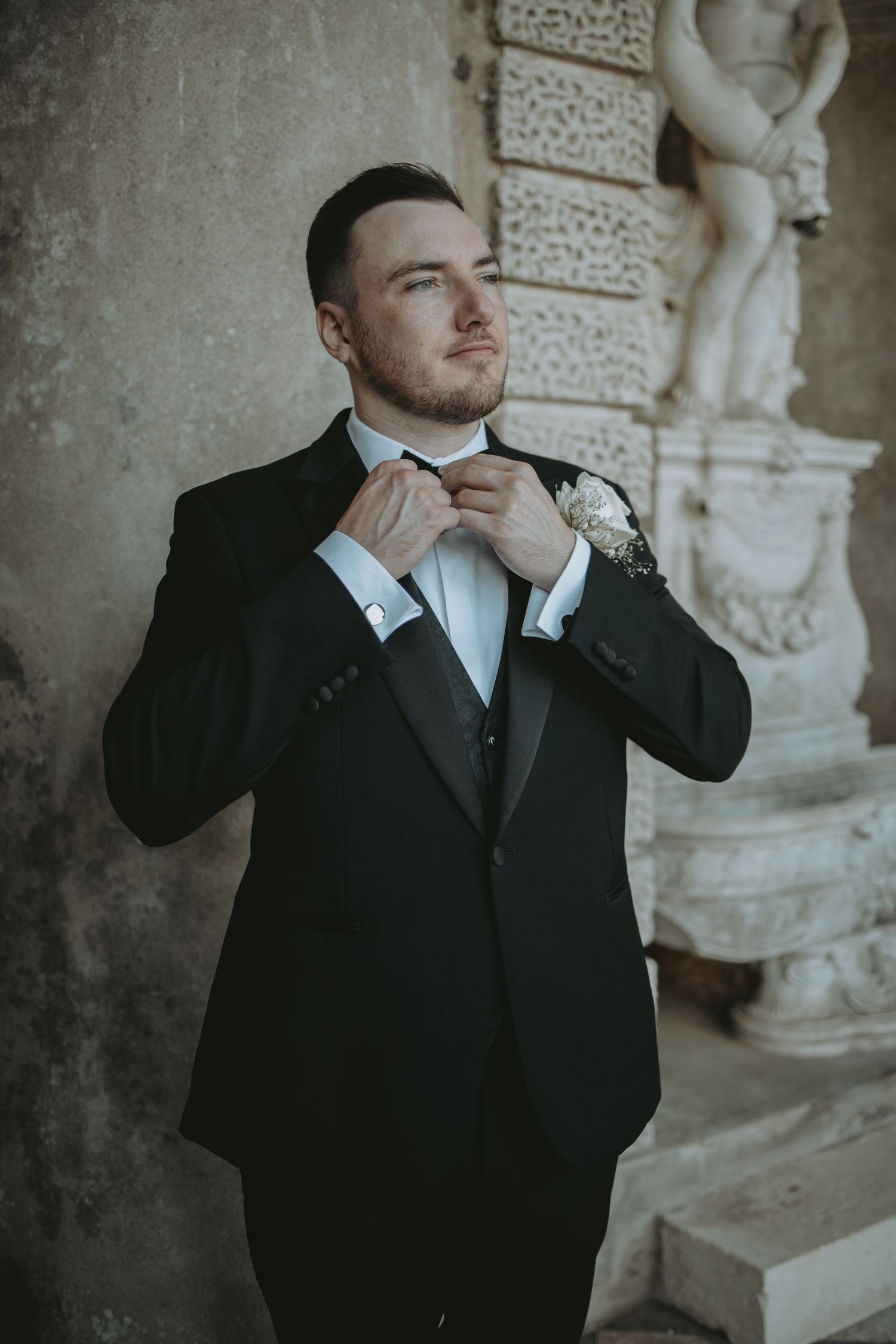 Groom in classic tuxedo poses against stone wall with decorative statue in background.