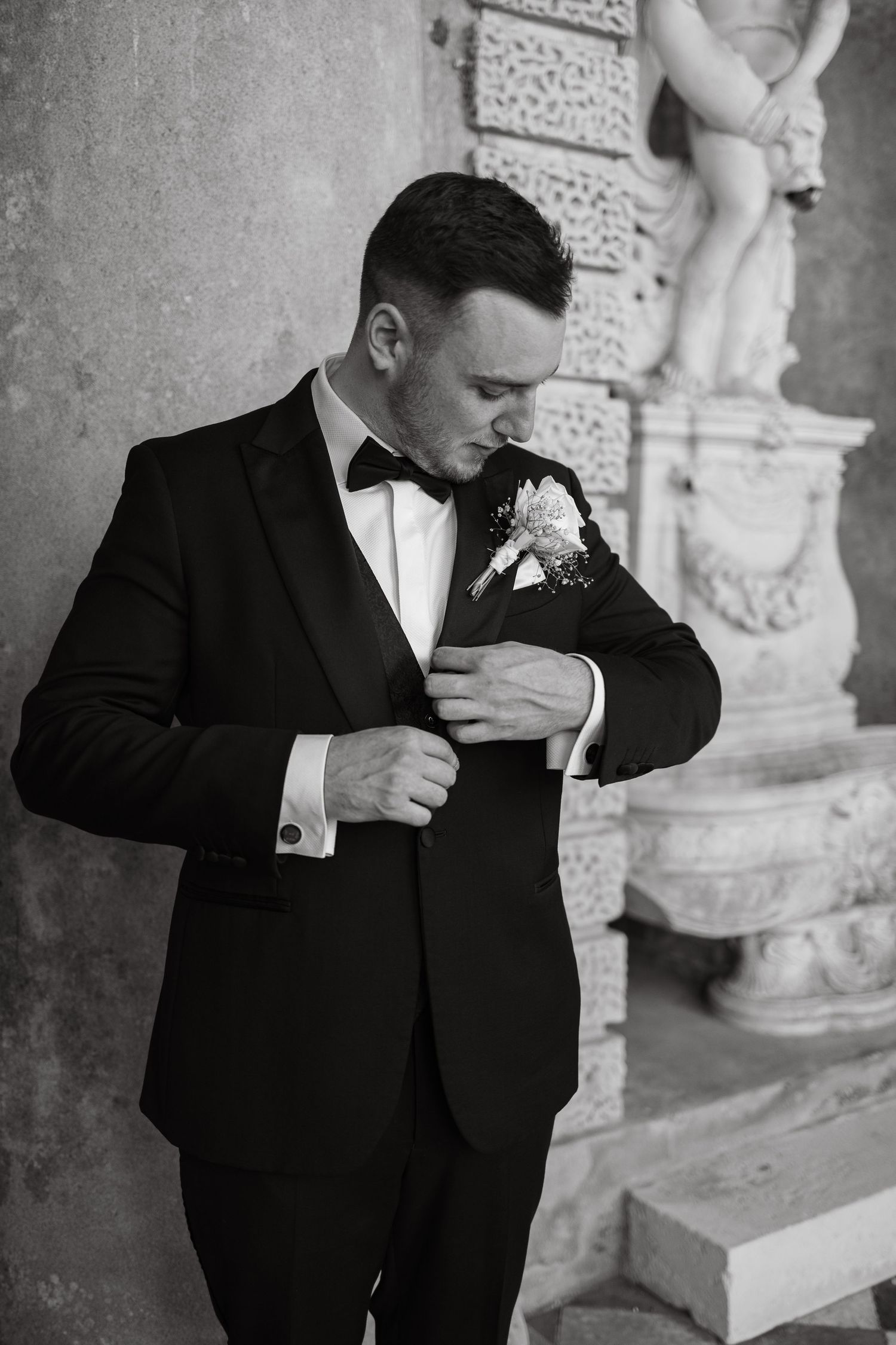 Groom adjusts his boutonniere while standing next to an ornate stone column.