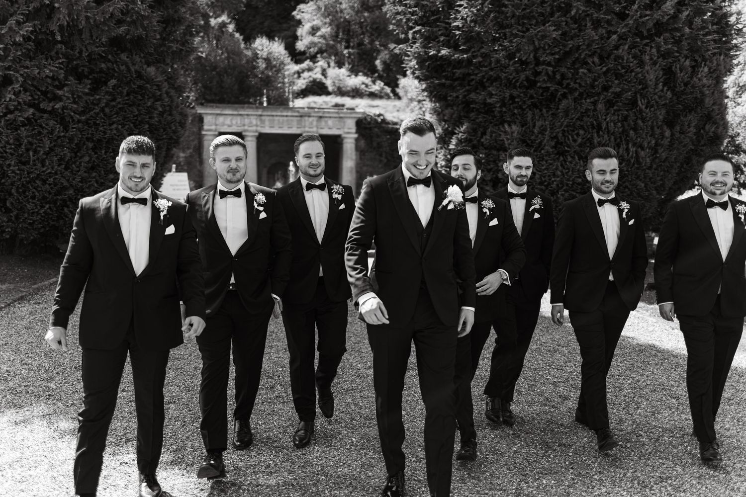 Row of groomsmen in black tuxedos walk together on grass during wedding day.