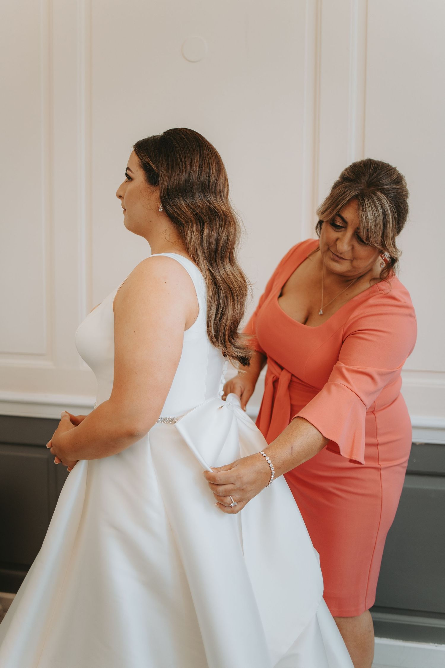 Person in coral dress helps adjust white wedding gown during bridal preparation.