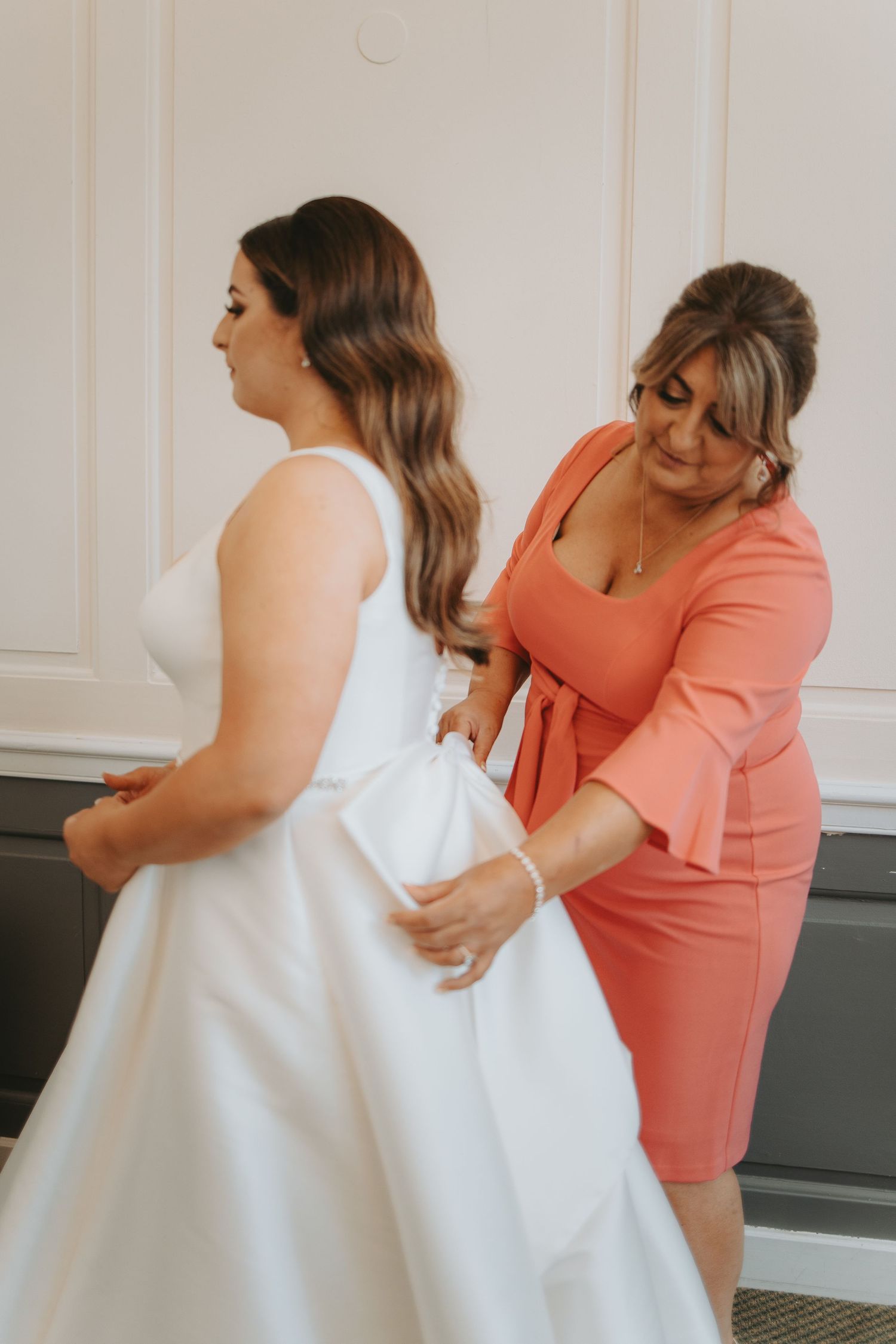 Helper in coral dress assists with fitting and adjusting white wedding dress.