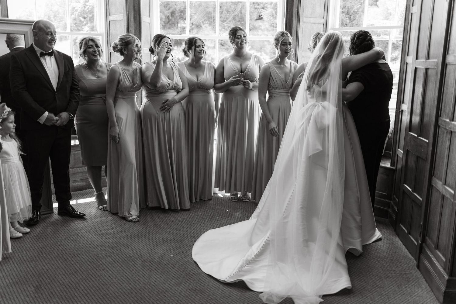 Wedding party gathers in elegant venue with large windows and formal attire.