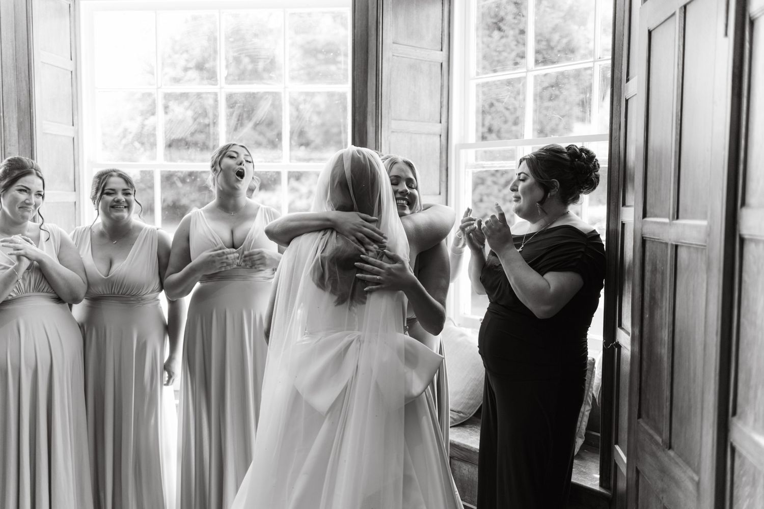 Wedding party members react emotionally in sunlit room with windows.