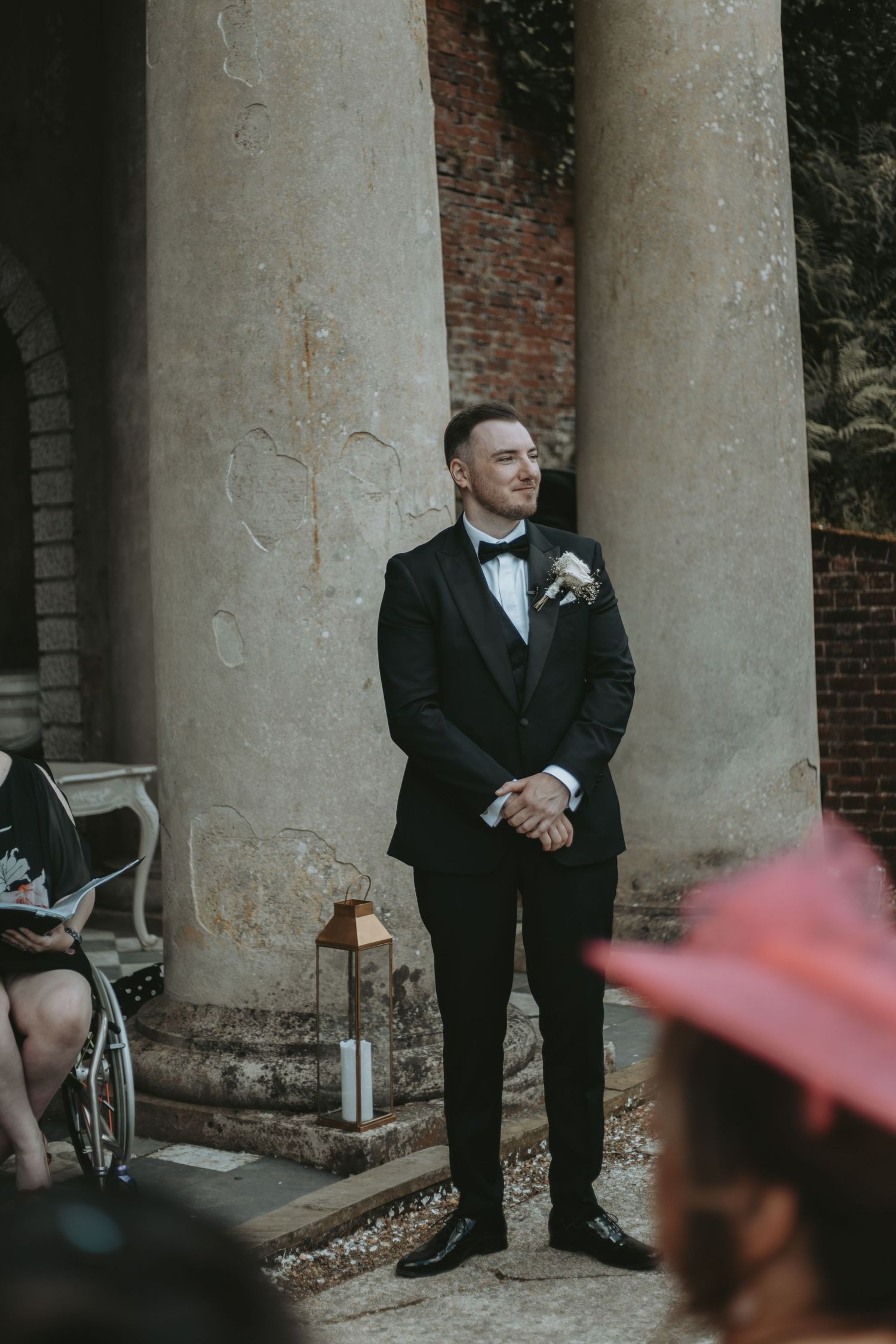 Formal wedding scene with people in black tuxedos standing between classical stone columns.