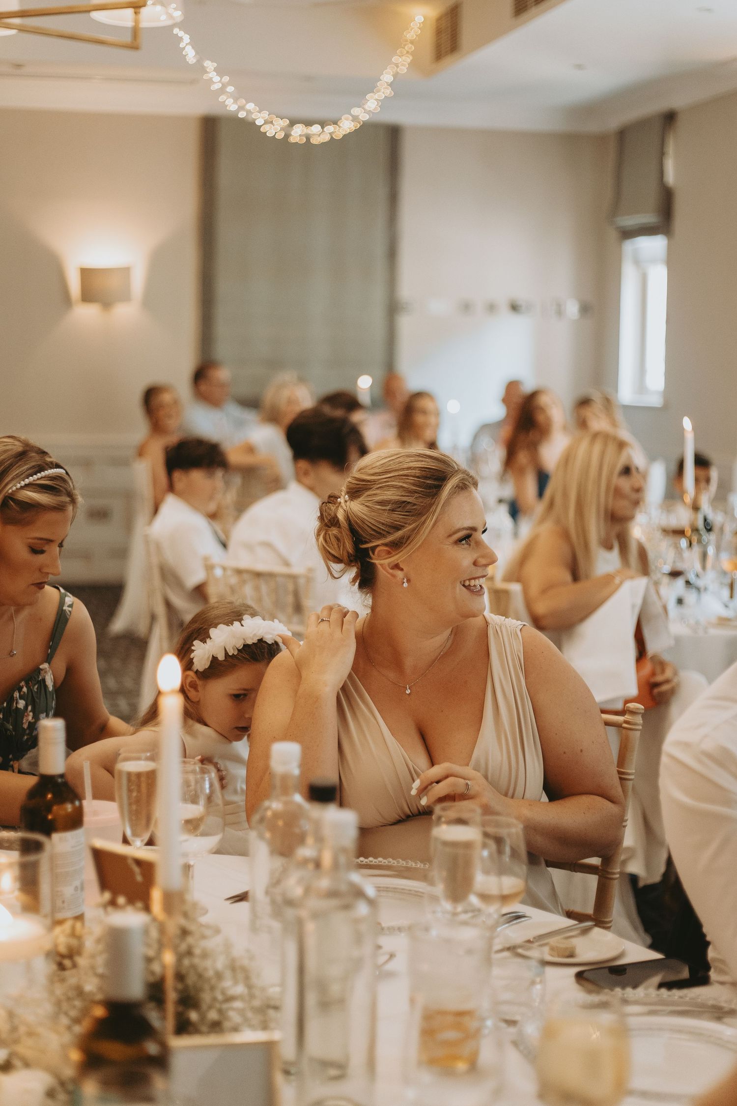Wedding guests laugh and smile during reception dinner at elegant white-decorated venue with candlelit tables.