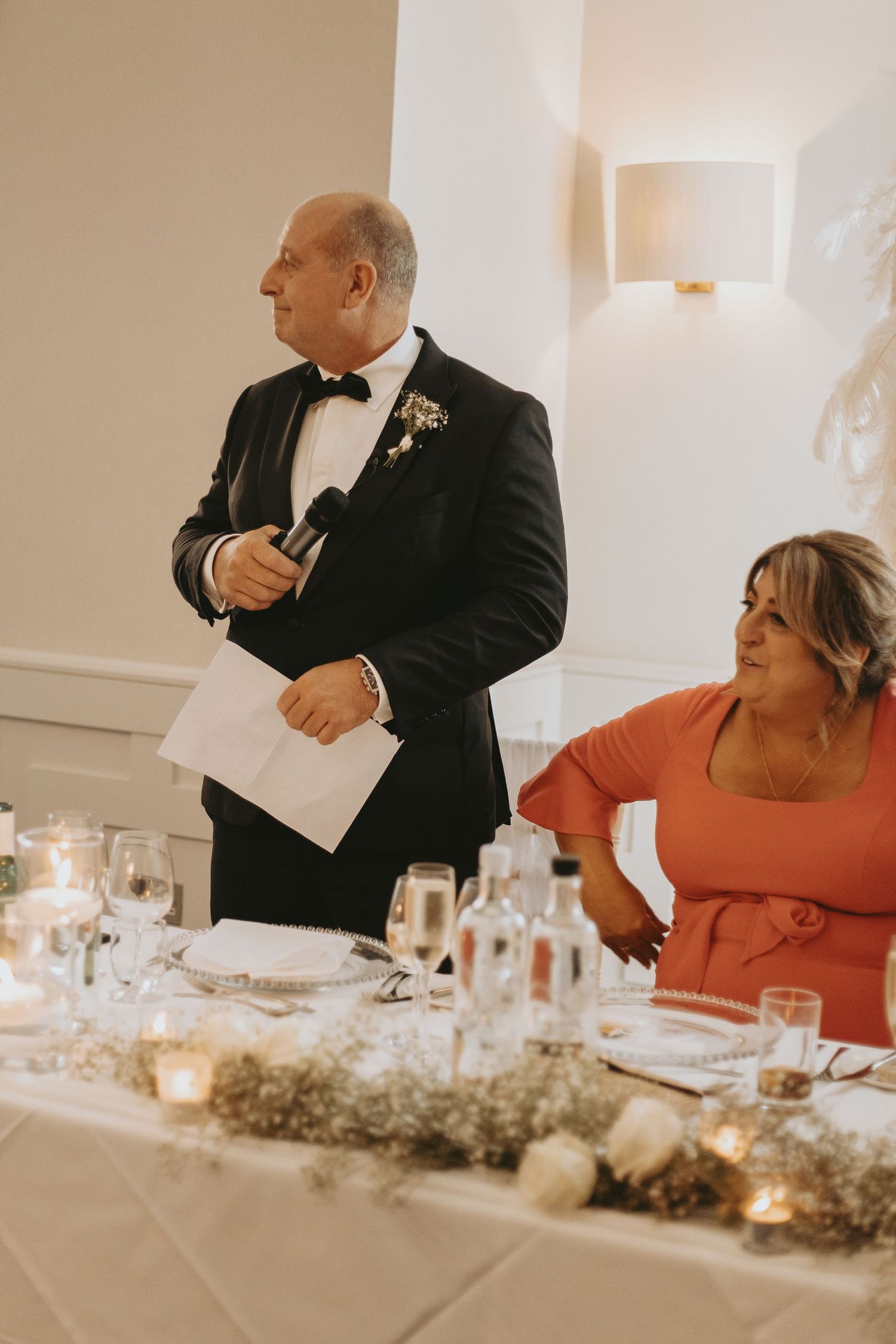Wedding reception guests seated at long table decorated with floral centerpieces and glowing candles.