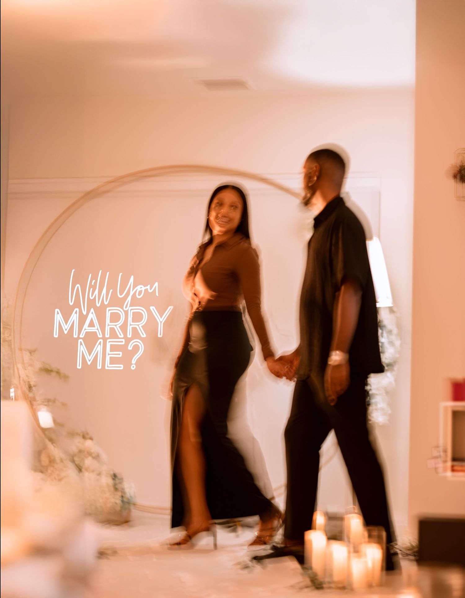 Two people stand in front of an illuminated 'Will You Marry Me?' sign with romantic candles creating an intimate atmosphere.