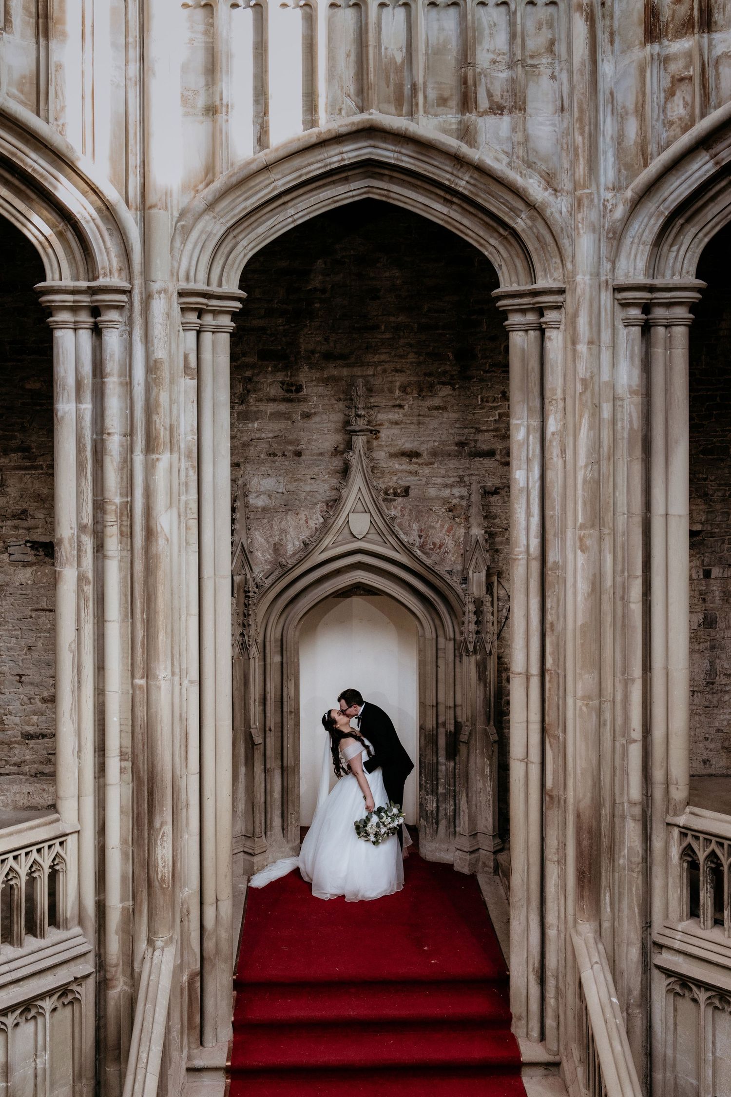 A couple stands in a Gothic stone archway with red carpet stairs in an elegant historic building interior at The Orangery Margam