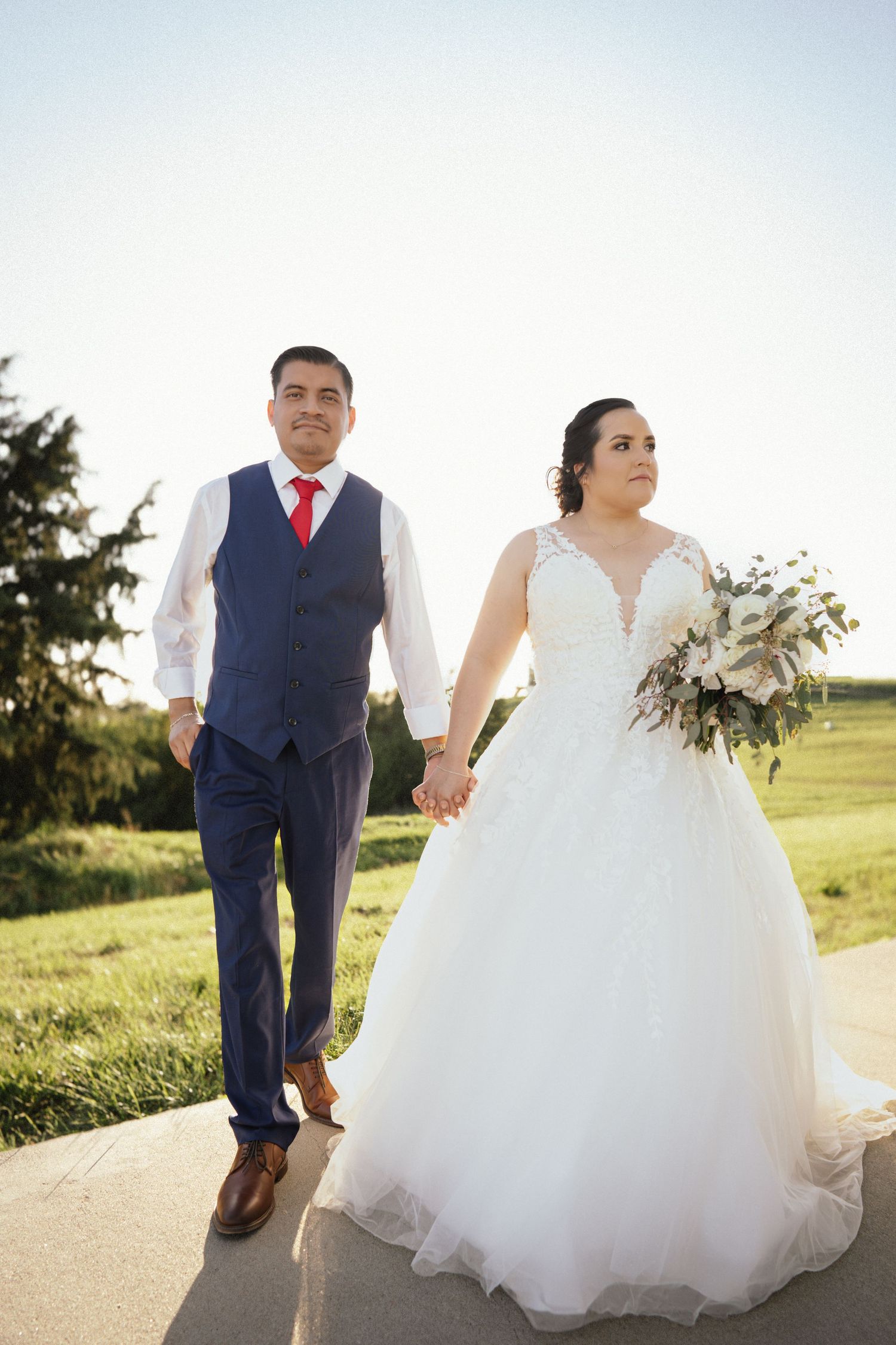 Wedding couple walking hand in hand on path with bride in lace gown and groom in navy vest.