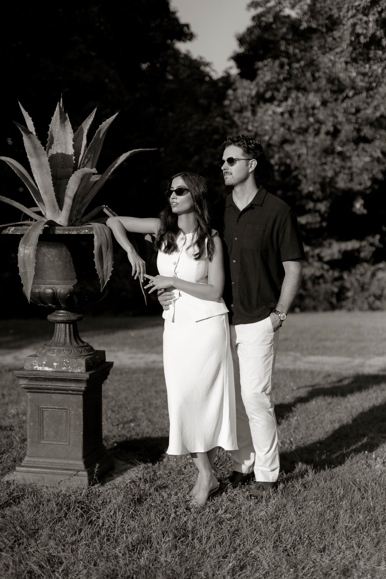 Black and white photo of couple standing by ornate planter in a garden setting.