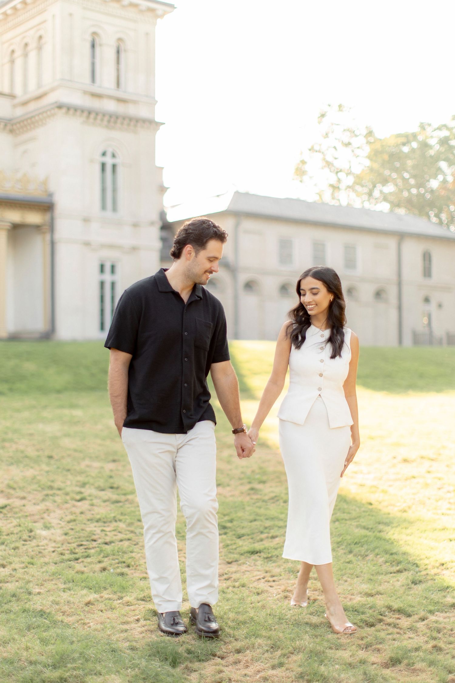 Elegant couple in coordinated outfits walking hand in hand near a church tower.