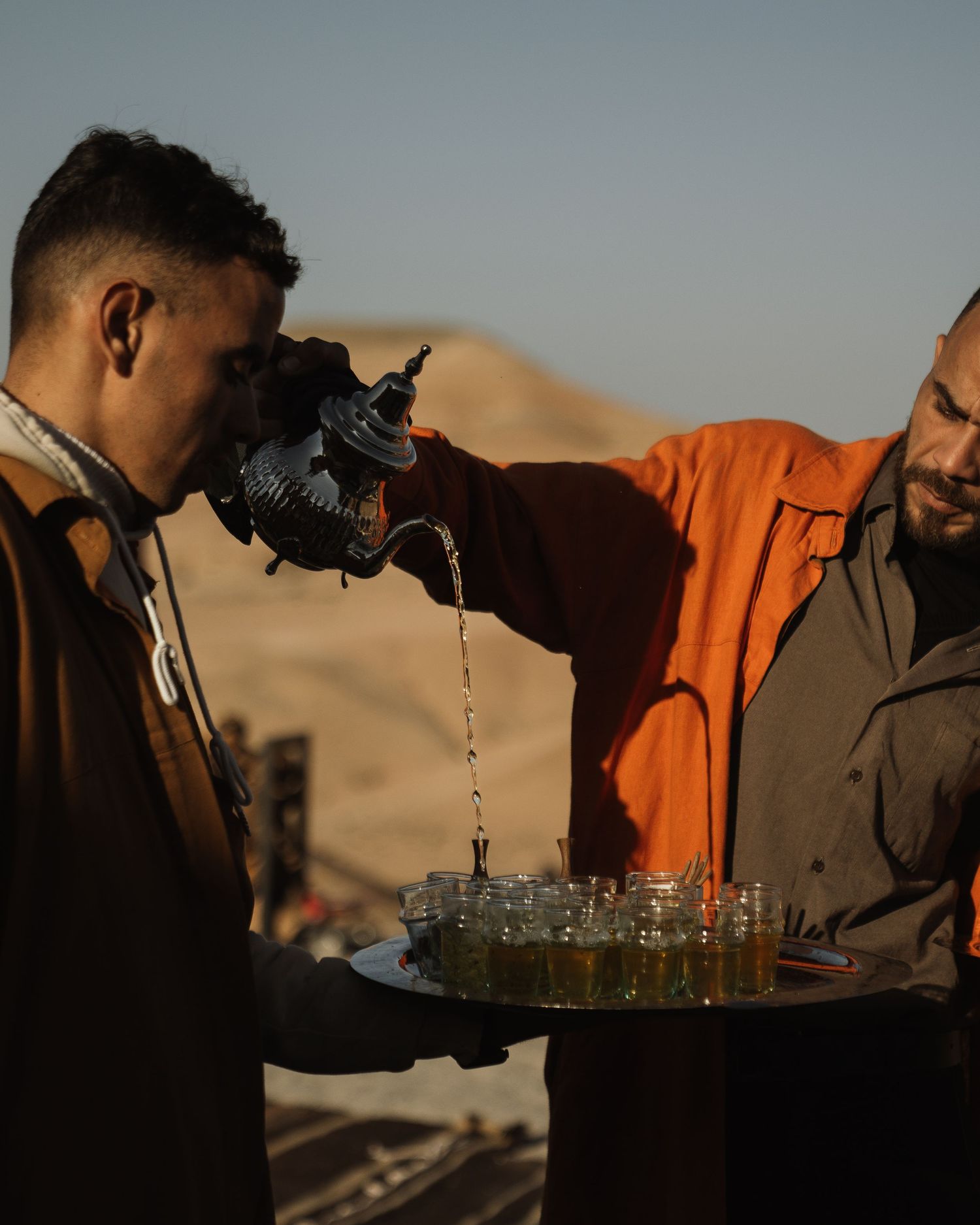 Tea being poured from traditional Moroccan teapot into small glasses at sunset.