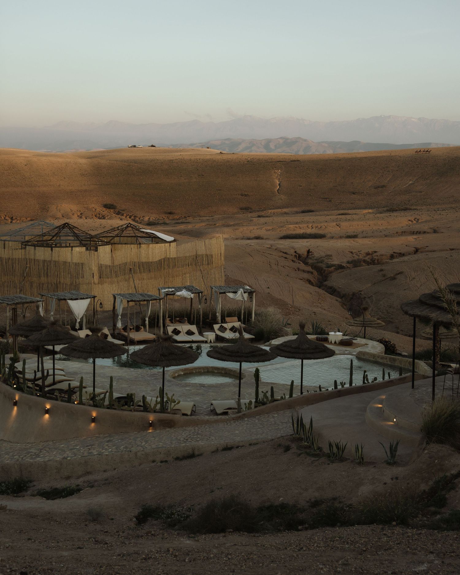 Luxury desert resort with illuminated pool and cabanas nestled among rolling sand dunes at dusk.