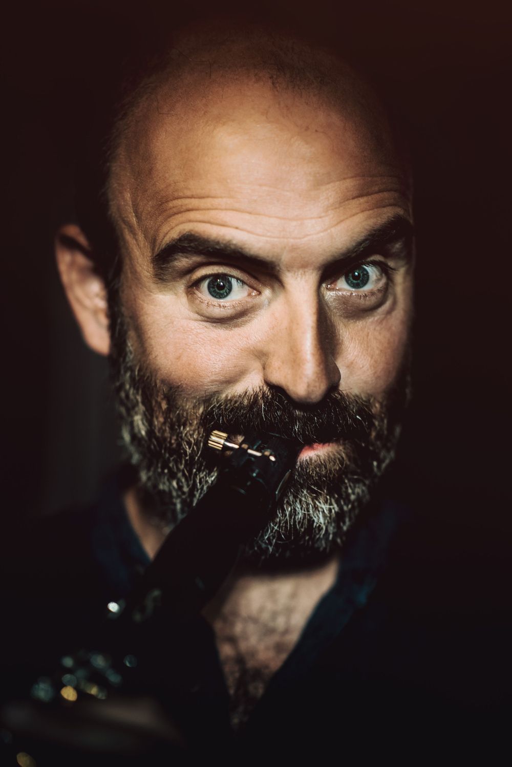 Dark artistic portrait of a bald musician with a grey beard playing a woodwind instrument in atmospheric lighting.