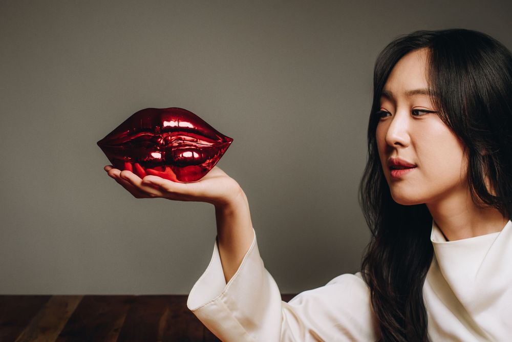 Side view of person in white sweater holding a large decorative red lip-shaped object against neutral background.