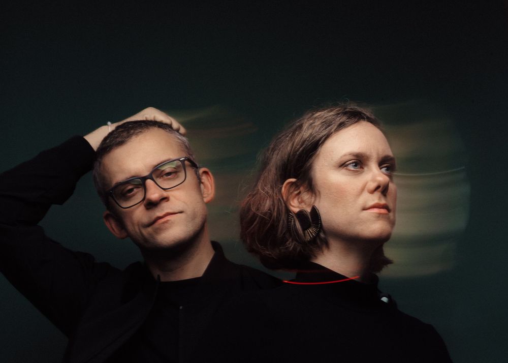 Portrait of two people in dark outfits against a dark background, one wearing glasses with hand on head.