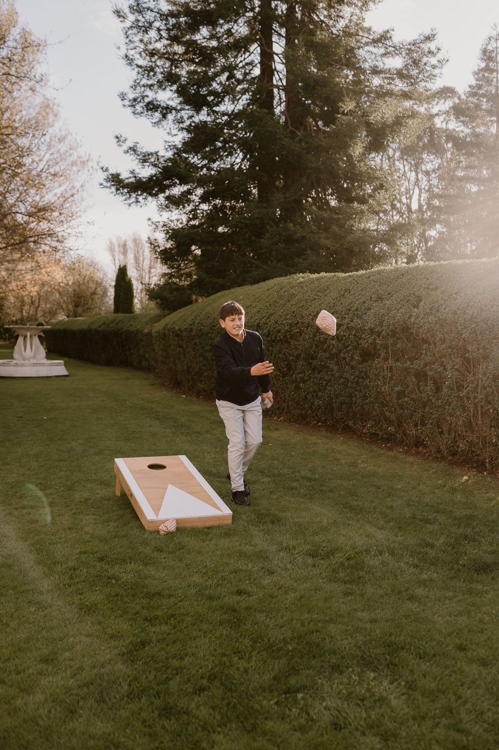 A wooden cornhole game board sits on a grassy lawn surrounded by trees and hedges in golden sunlight.