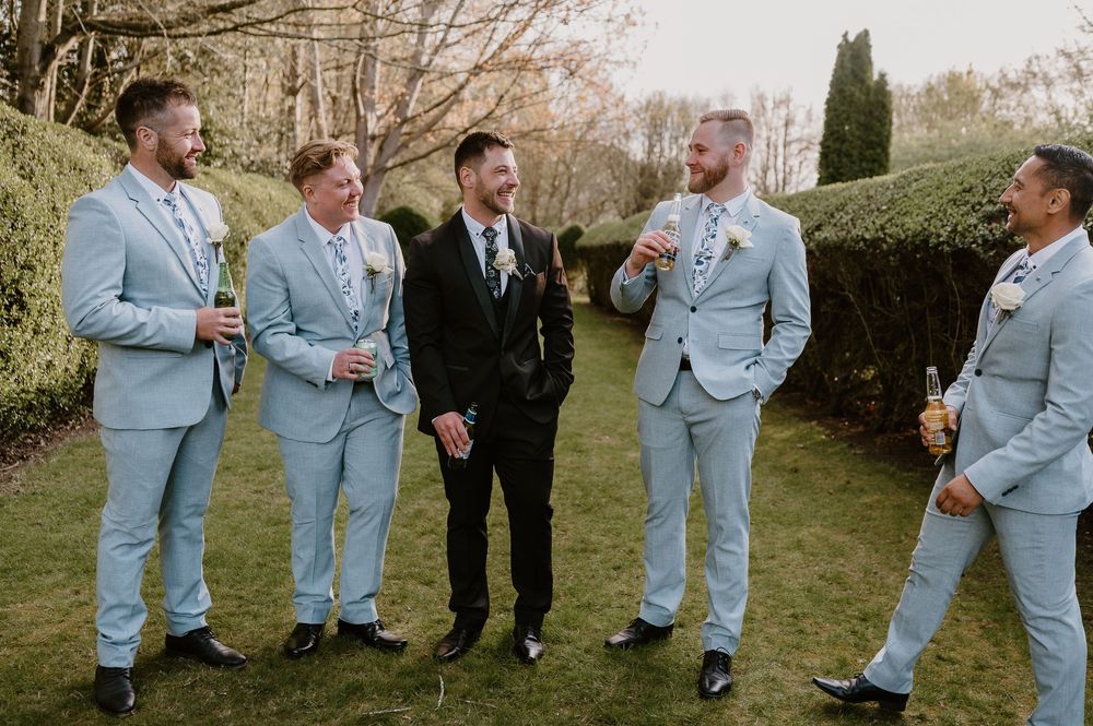 Wedding party in light blue suits laughing together on a sunny afternoon outdoors.