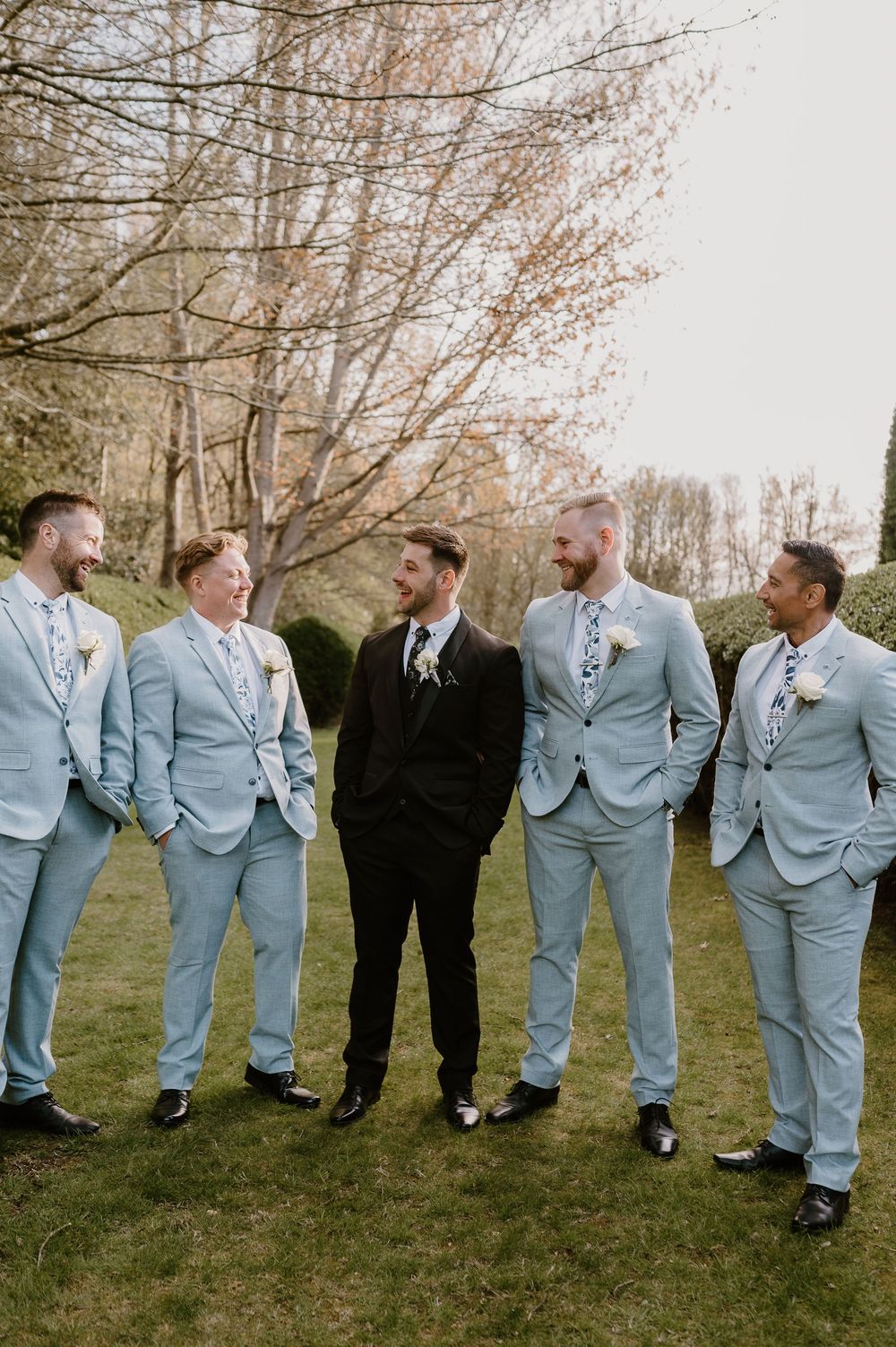 Groomsmen in light blue suits gather outdoors with the groom in black, sharing a candid moment on the lawn.