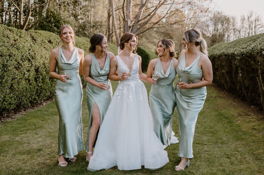 A bridal party in matching sage green bridesmaid dresses poses on a garden lawn with the bride in a white gown.