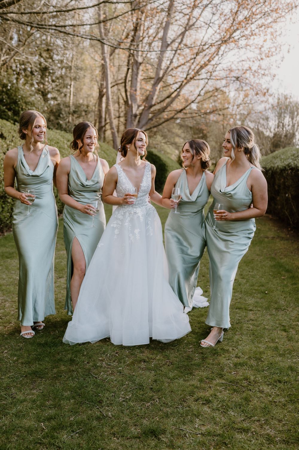 Wedding party wearing sage green bridesmaid dresses and white bridal gown walking together on grass in outdoor setting.