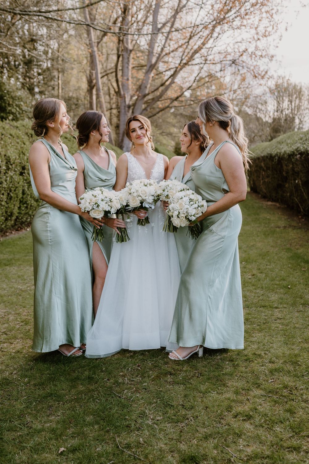 Bridesmaids in matching sage green dresses pose with bouquets of white flowers in a garden setting.