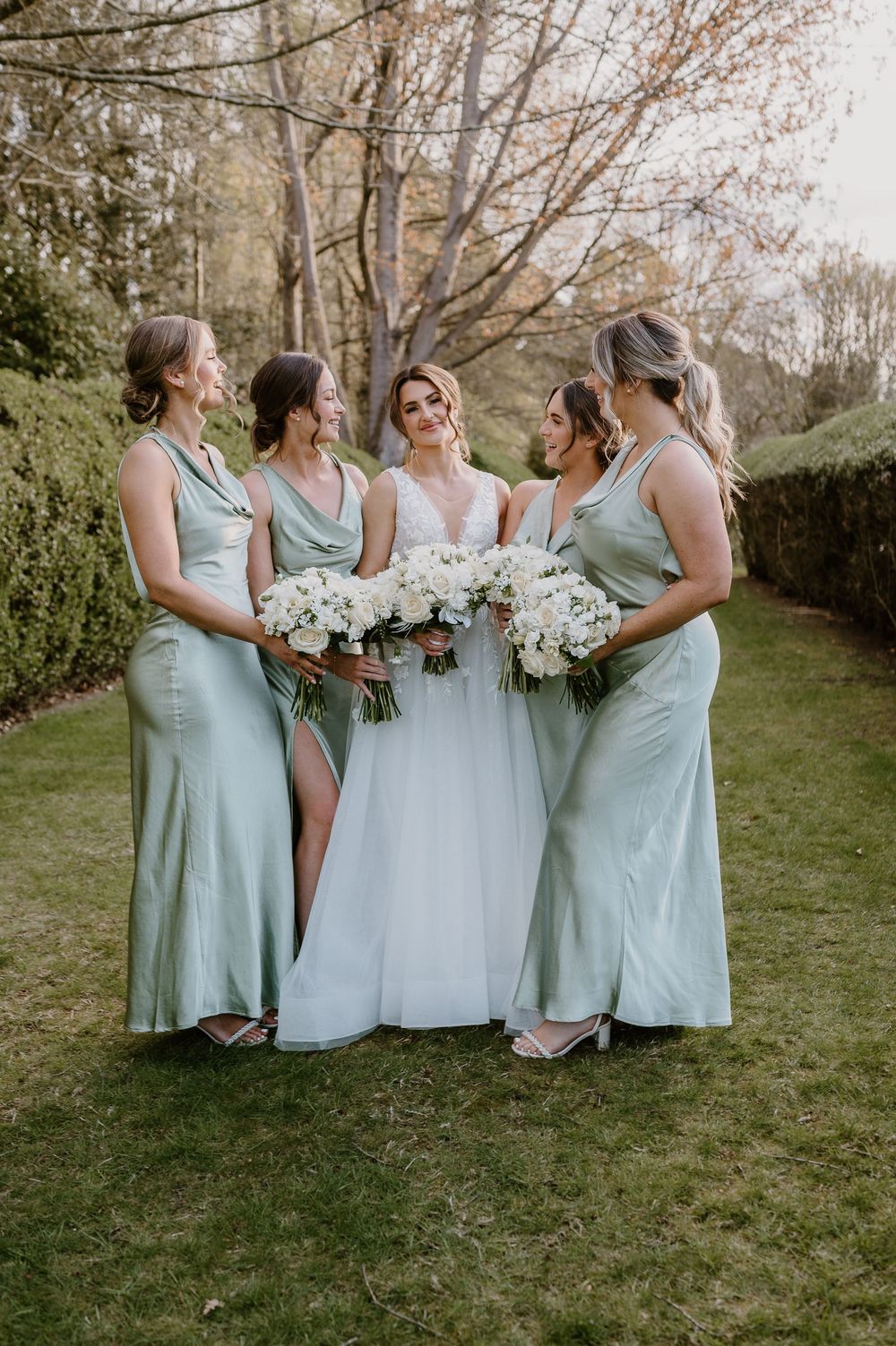 A wedding party in sage green bridesmaid dresses poses together on a grassy lawn holding white bouquets.