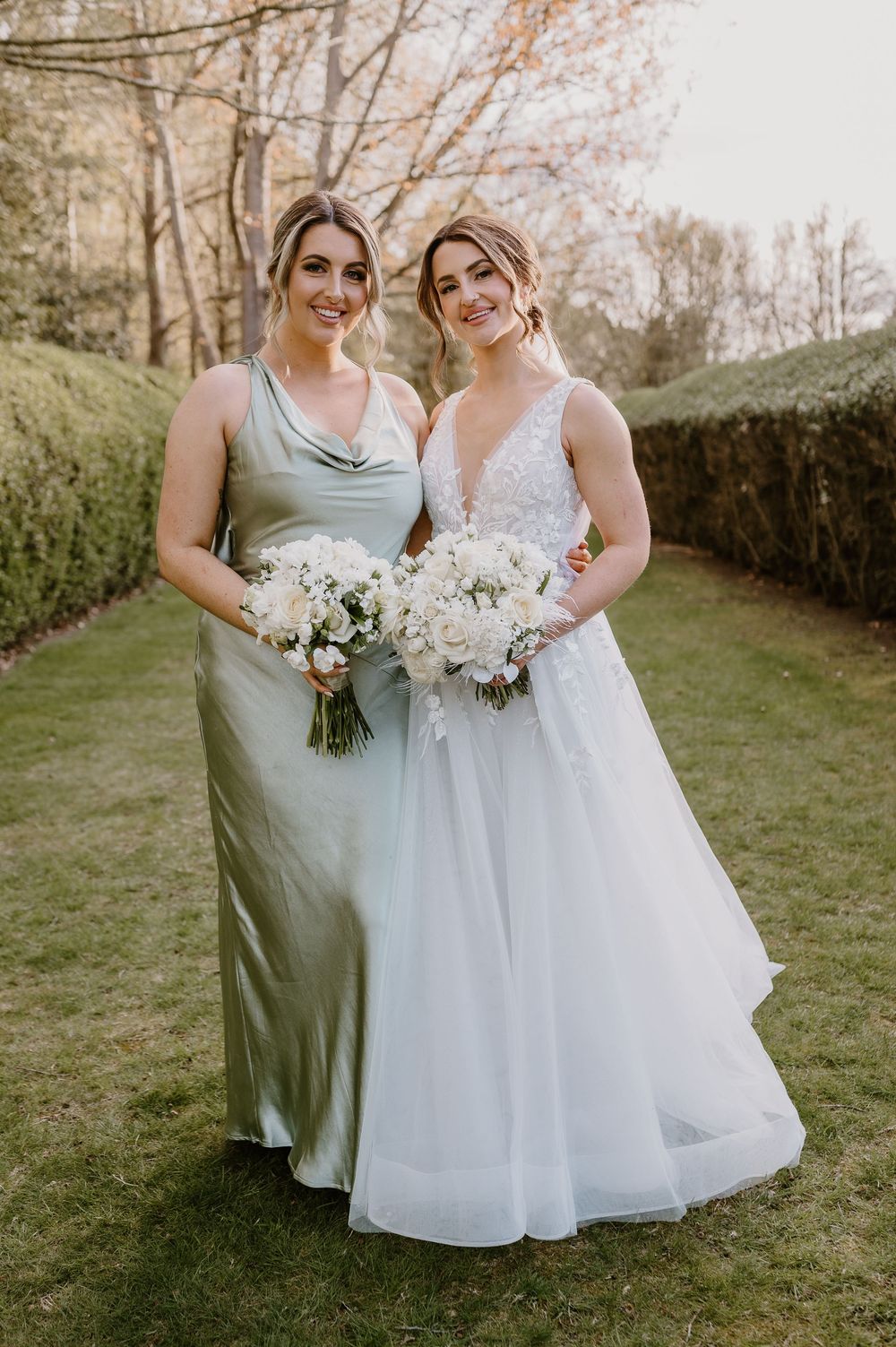 Wedding party members in sage green and white dresses pose with bouquets in a garden setting.