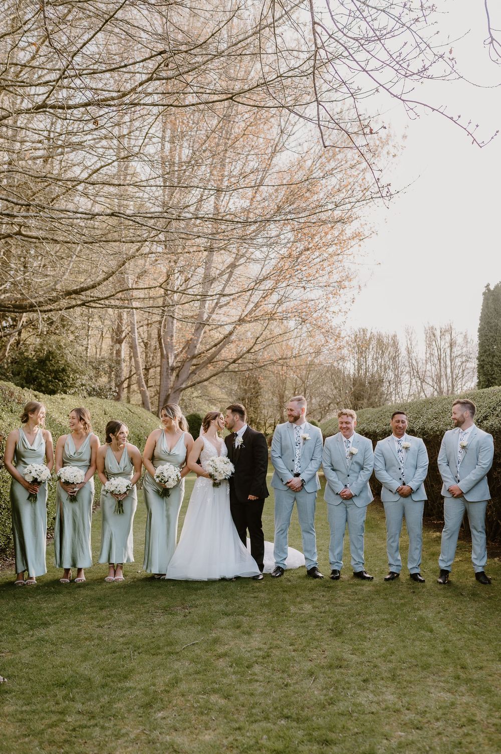 Wedding party wearing light blue and white stands in line against bare trees in outdoor garden setting during golden hour.