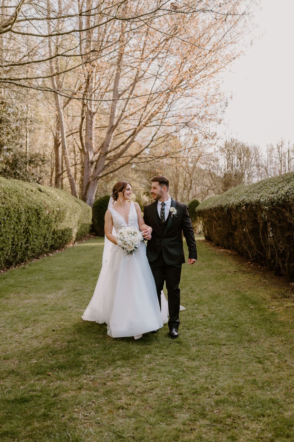 A wedding couple walks through a picturesque garden path lined with bare trees on a spring day.