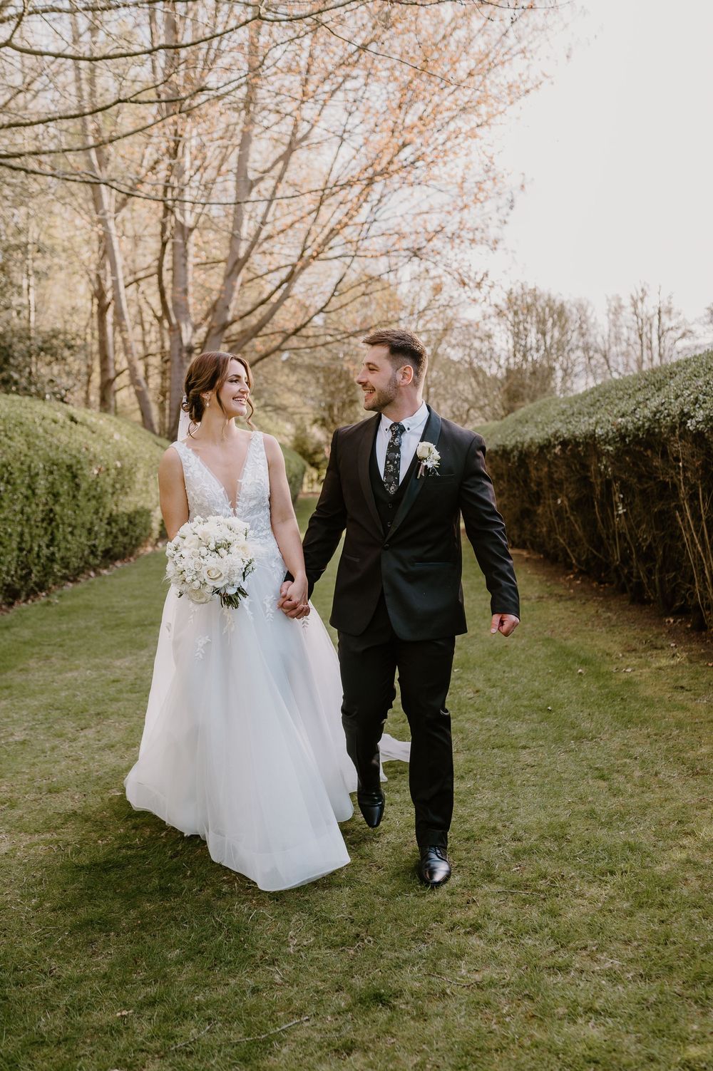 A bride and groom walk hand in hand along a hedge-lined garden path during their wedding.