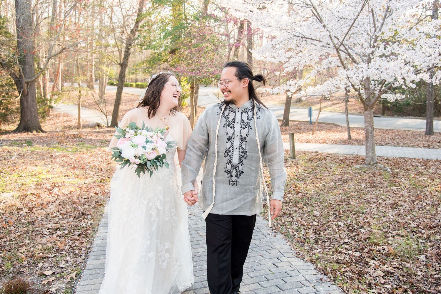 A couple walking hand in hand through a scenic wooded area with fallen autumn leaves on the ground.