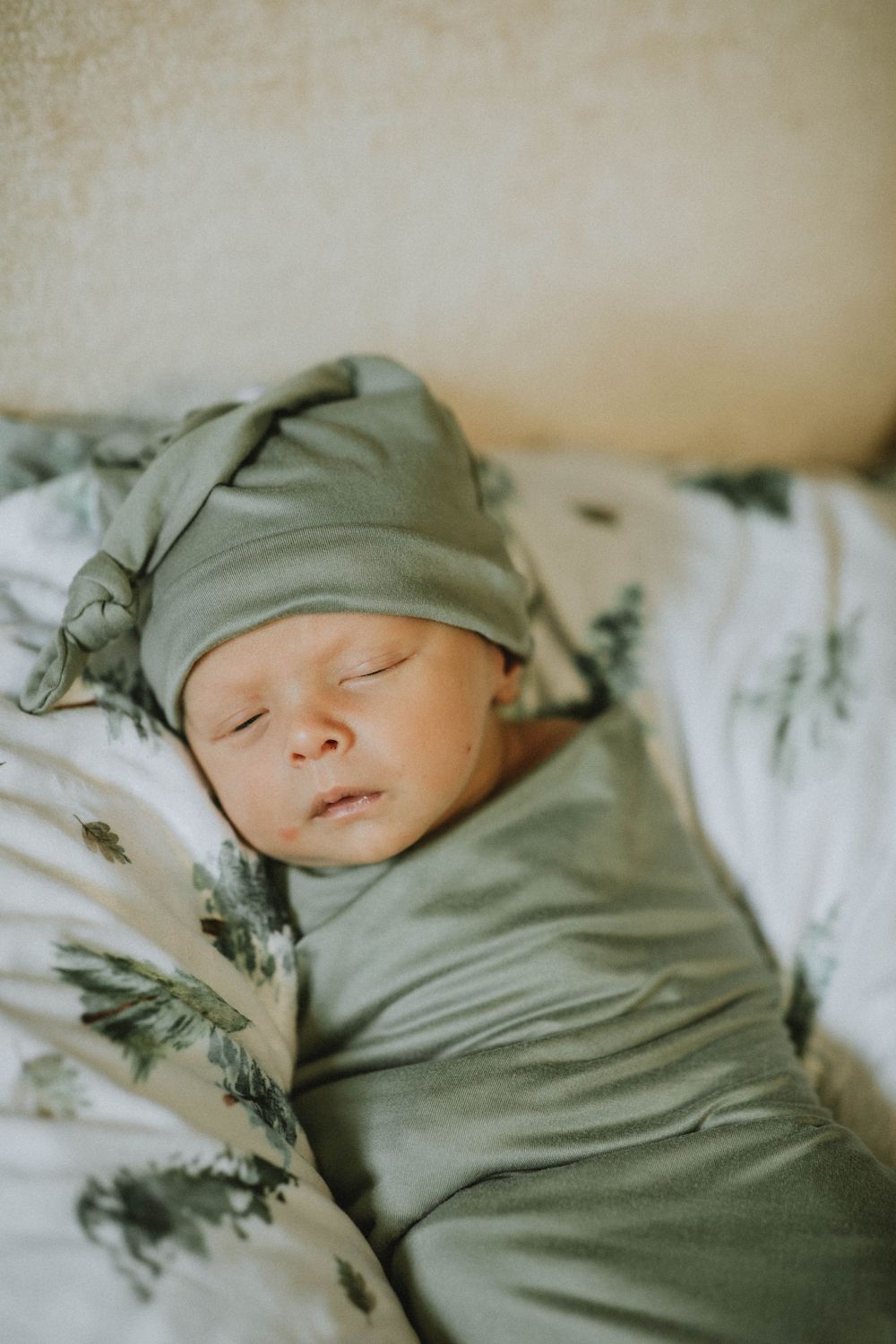 A sleeping newborn baby swaddled in soft sage green fabric rests peacefully on white bedding.