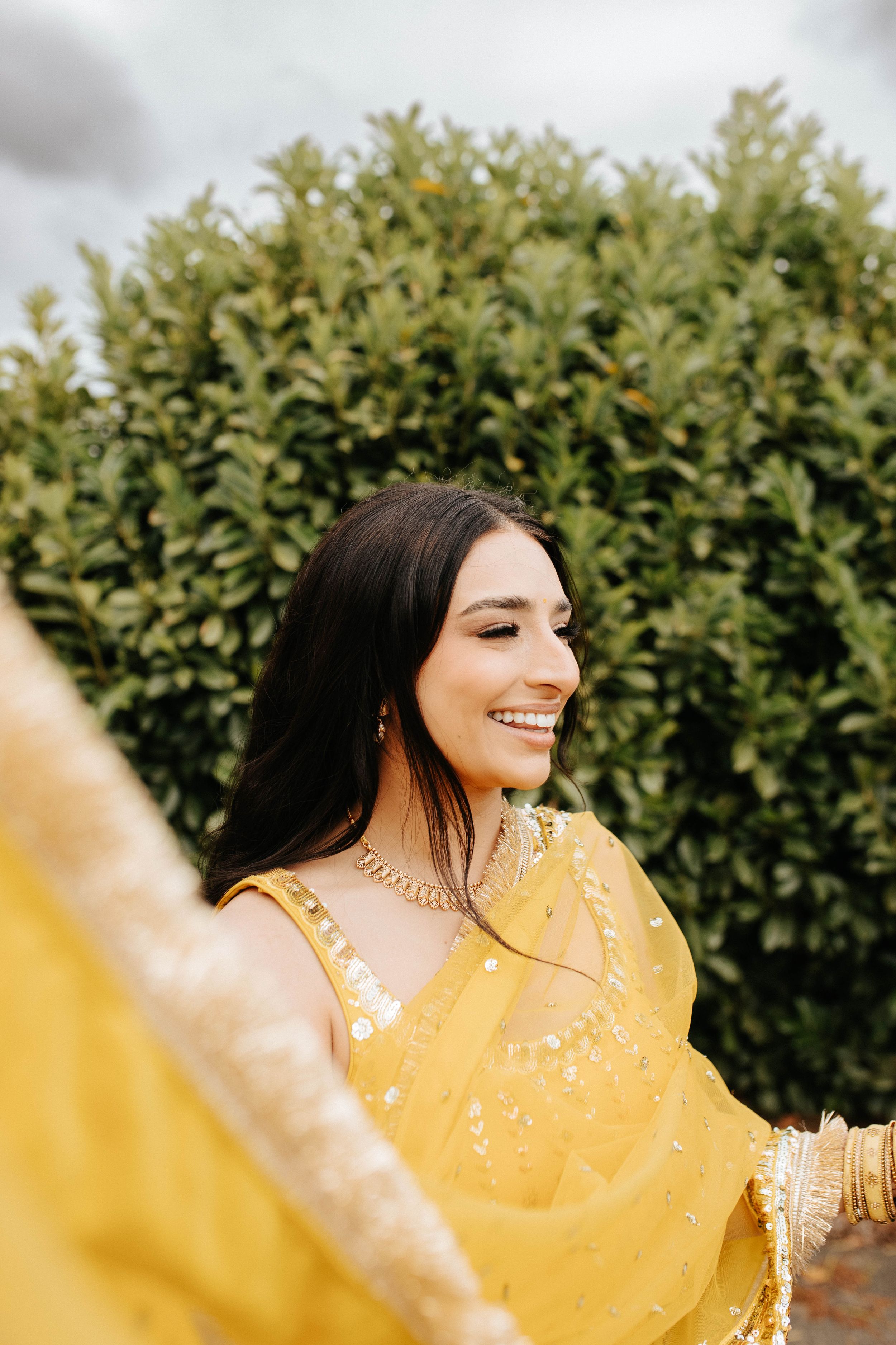 A person wearing a vibrant yellow traditional Indian sari smiles brightly against green foliage.