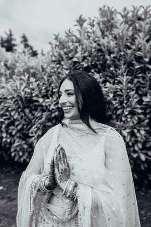 Black and white portrait of someone in traditional South Asian attire with henna-decorated hands smiling outdoors.