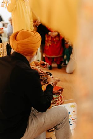 Person wearing orange turban seated during traditional Sikh ceremony with decorative items nearby.