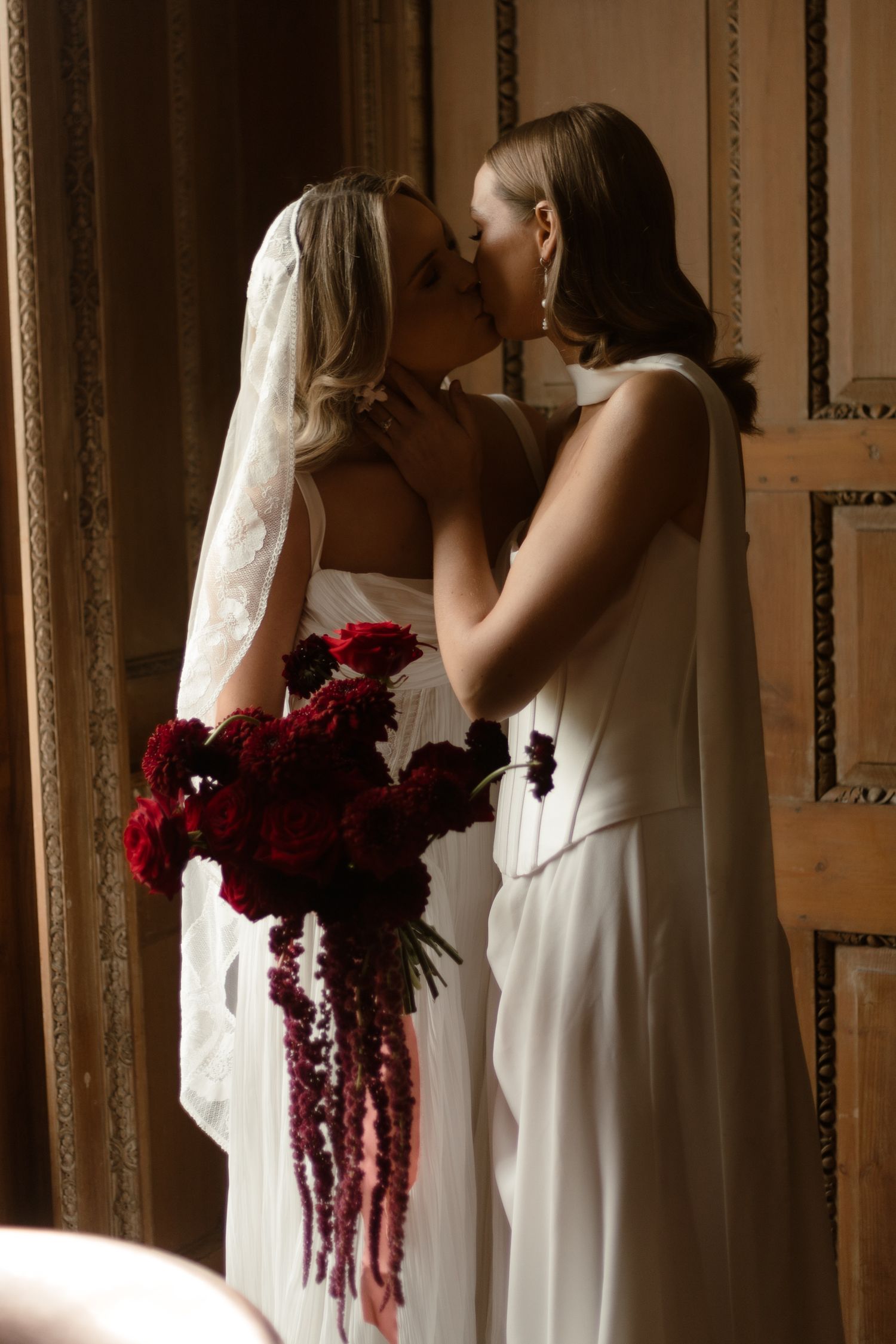 Two people share a romantic wedding kiss while holding a dramatic cascading bouquet of deep red flowers.