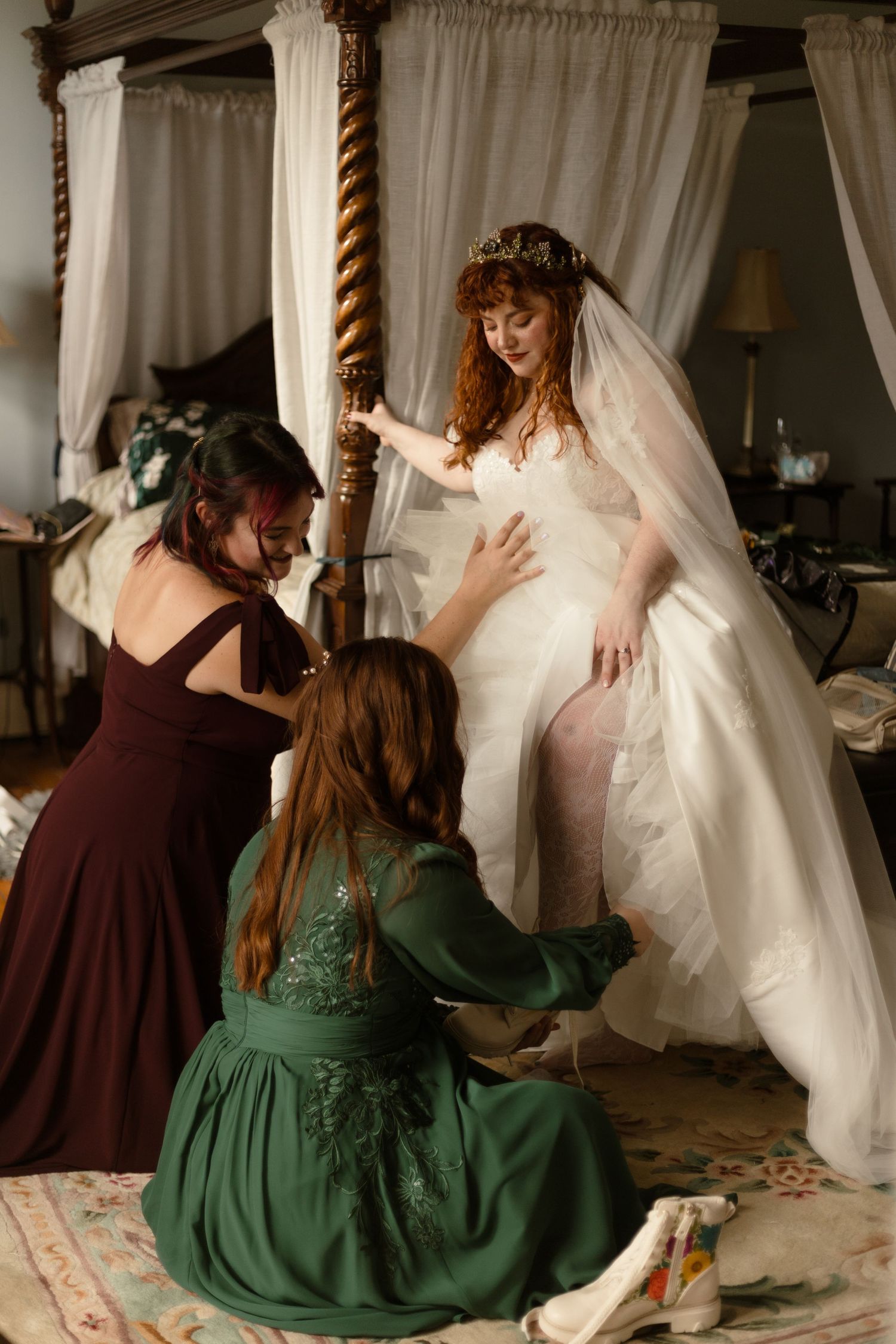 Two people in contrasting brown and emerald dresses help adjust a flowing white wedding gown in an intimate setting.