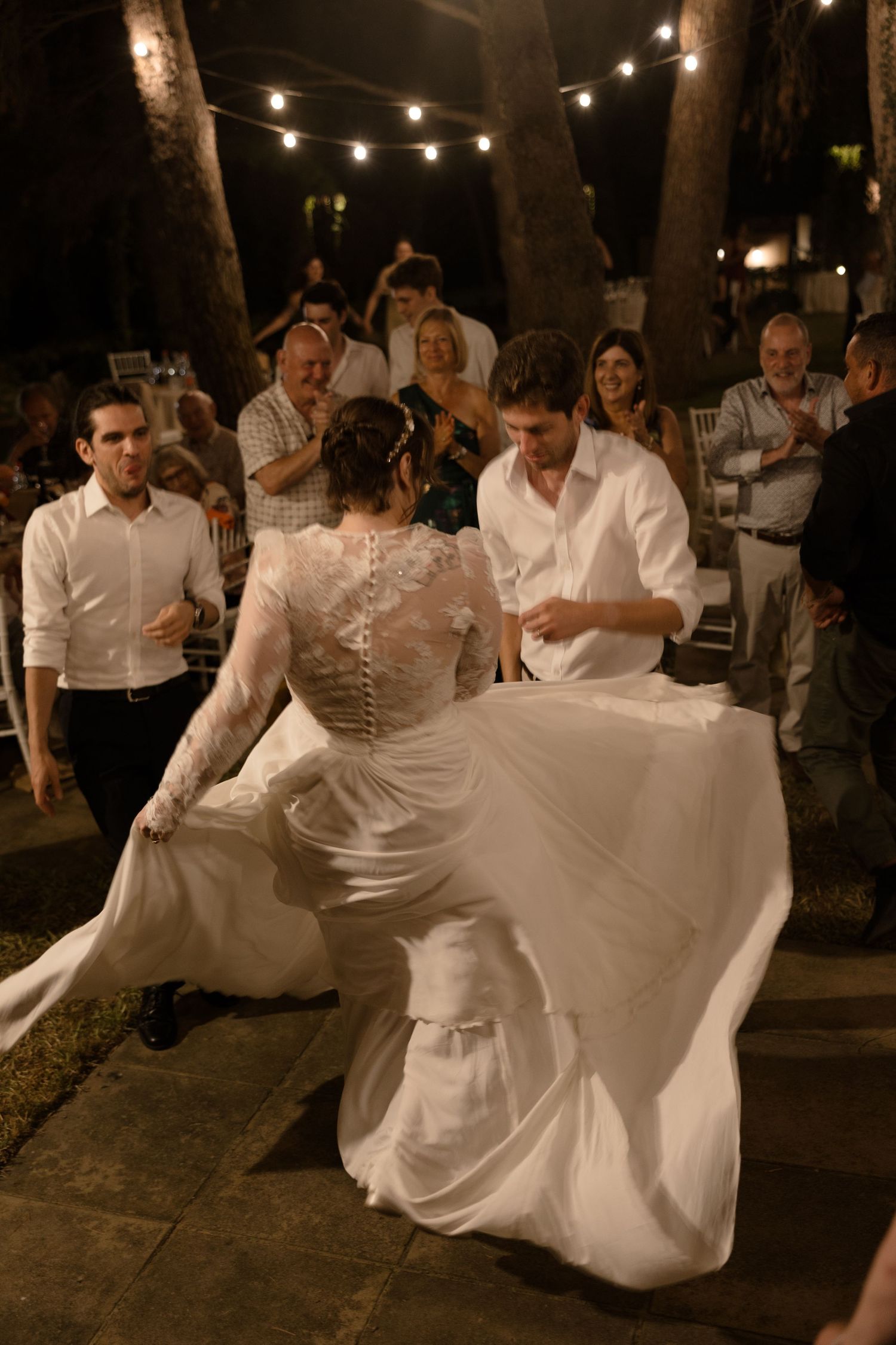 A couple dances at a nighttime outdoor wedding celebration with string lights overhead as guests watch.