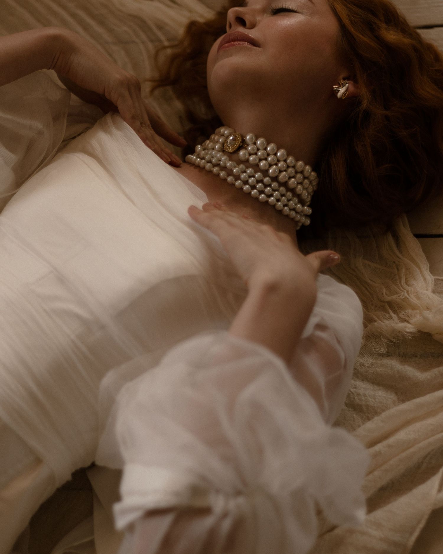 Figure lounging in dreamy lighting wearing pearls and white dress against cream colored bedding.