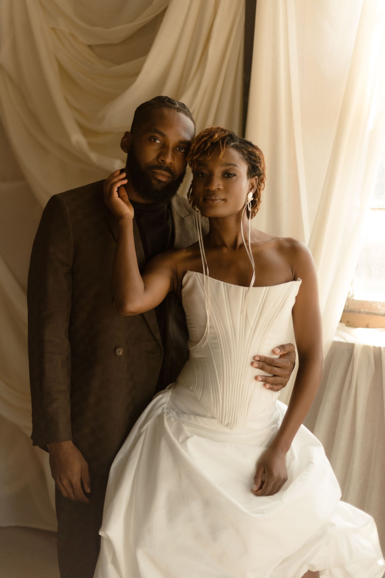 A couple poses together against white drapes, with the bride wearing a white corset wedding gown and the groom in a brown suit.