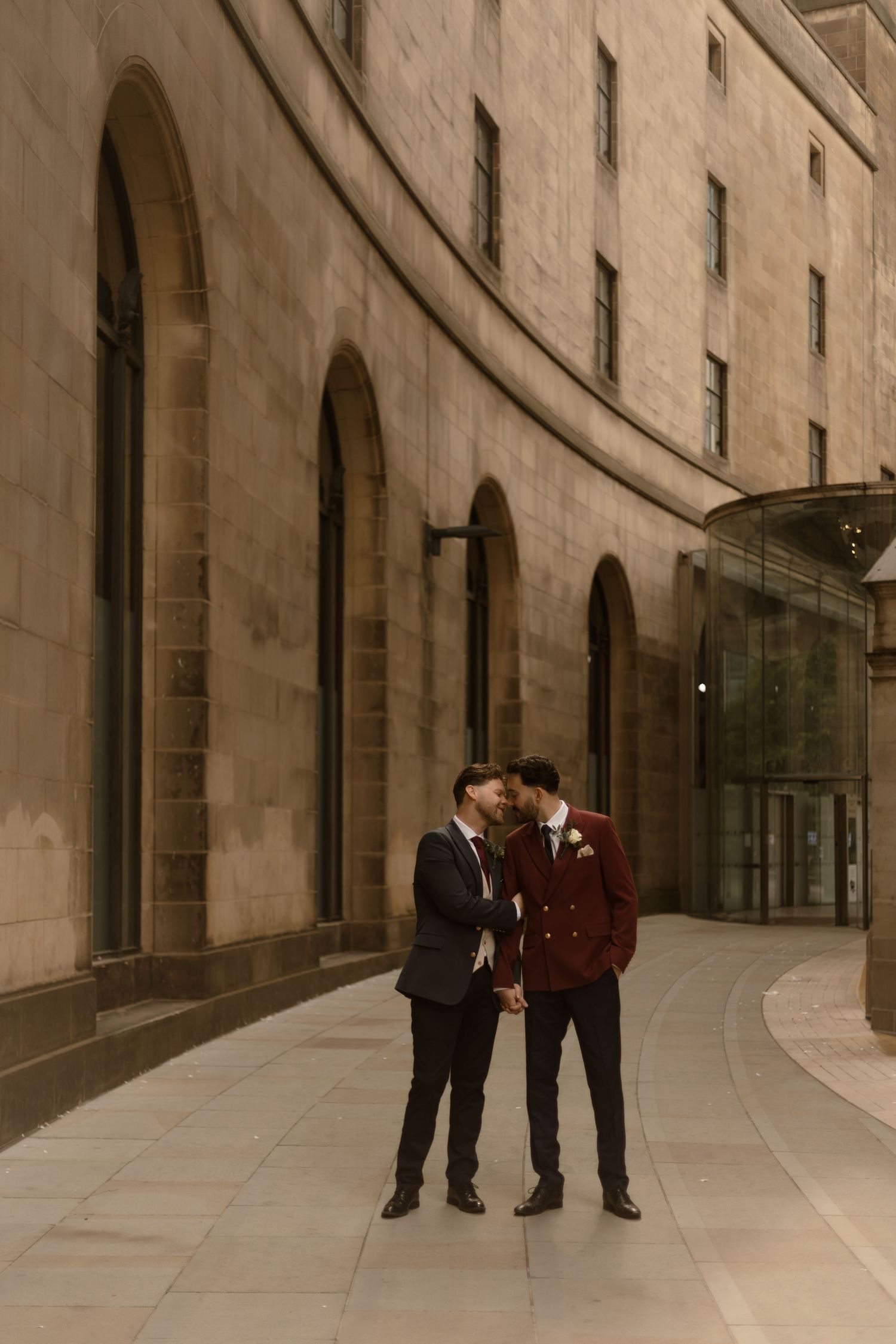 Two people in formal attire share an intimate moment against a curved stone building with arched windows.