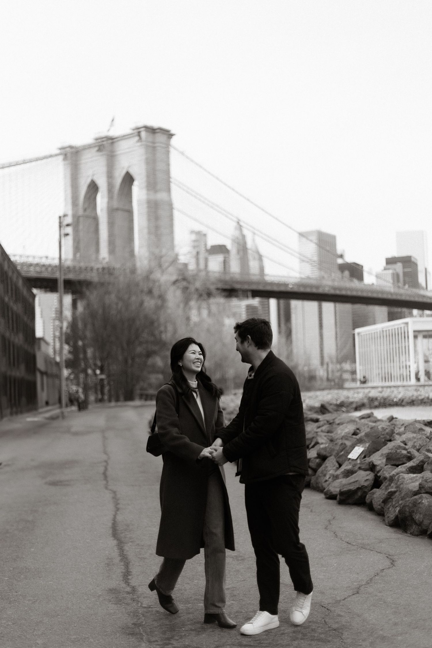 Black and white photo series of two women walking and talking along the Brooklyn Bridge waterfront path with bridge towers visible.
