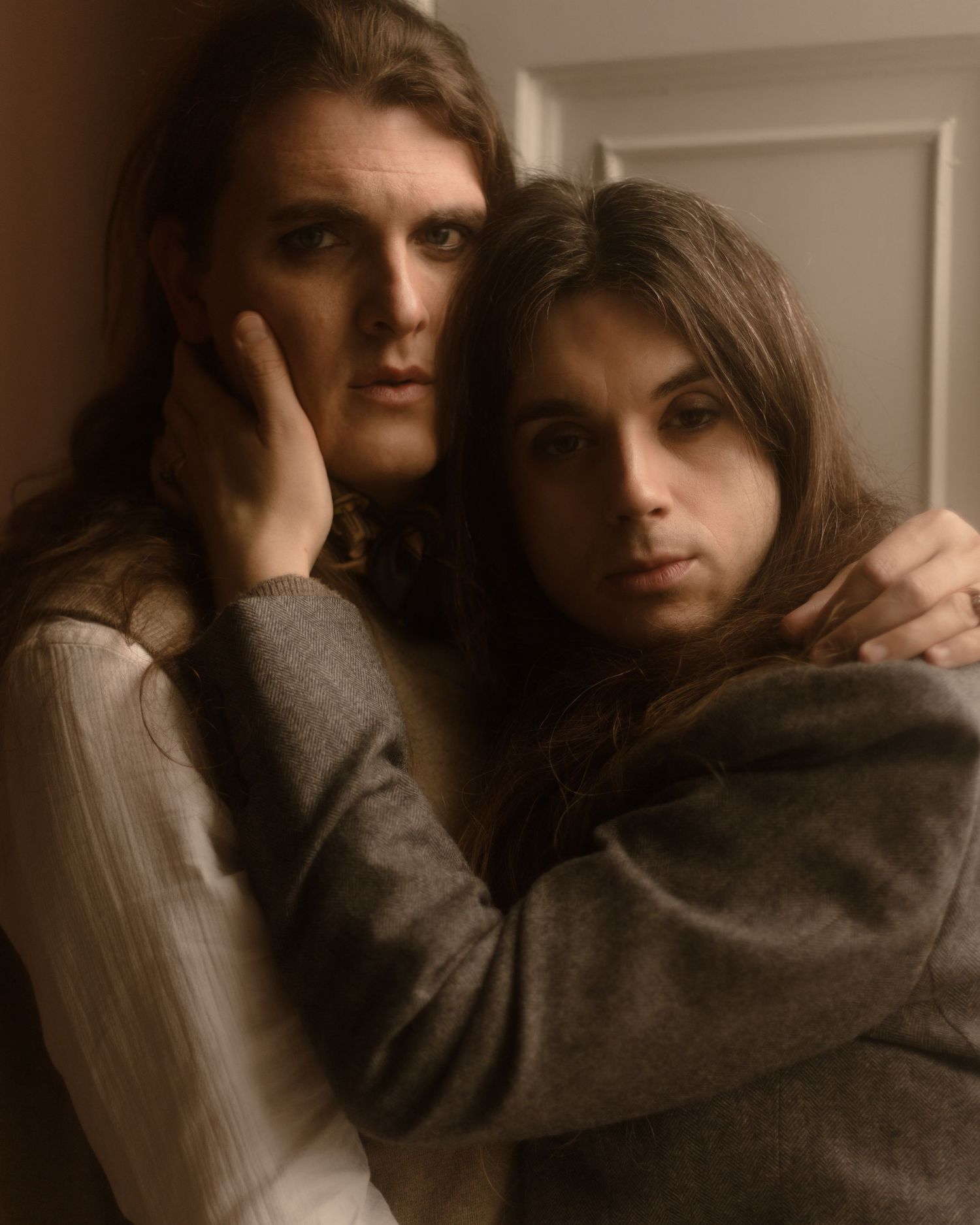 A moody, sepia-toned portrait of two people in intimate embrace against a light colored wall.
