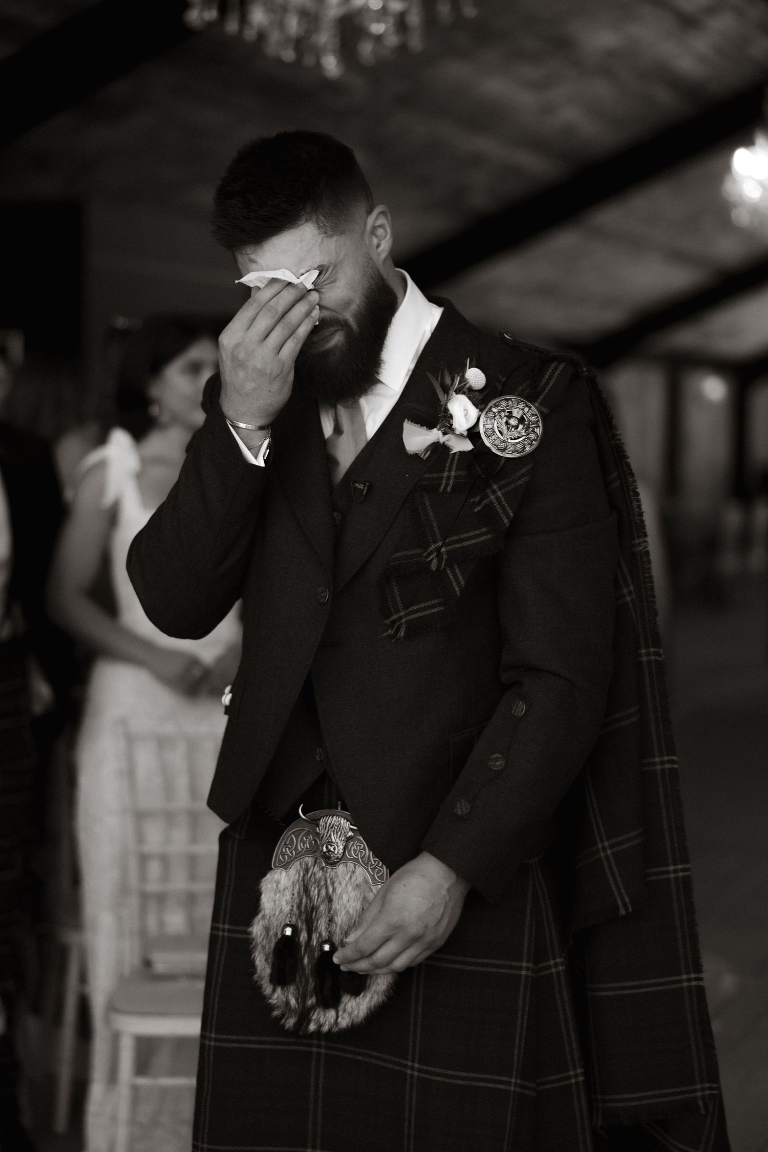 Black and white photo showing an emotional moment of a groom in traditional Scottish kilt attire at a wedding ceremony.