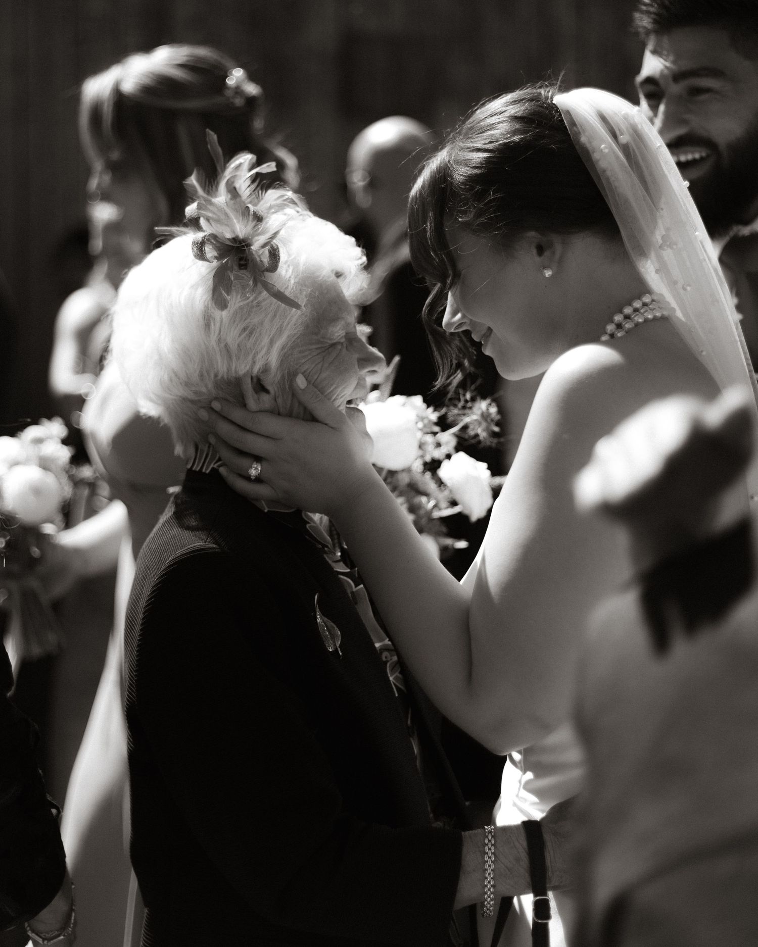 Black and white wedding photo showing a tender moment between figures as they embrace and share laughter at an outdoor celebration.