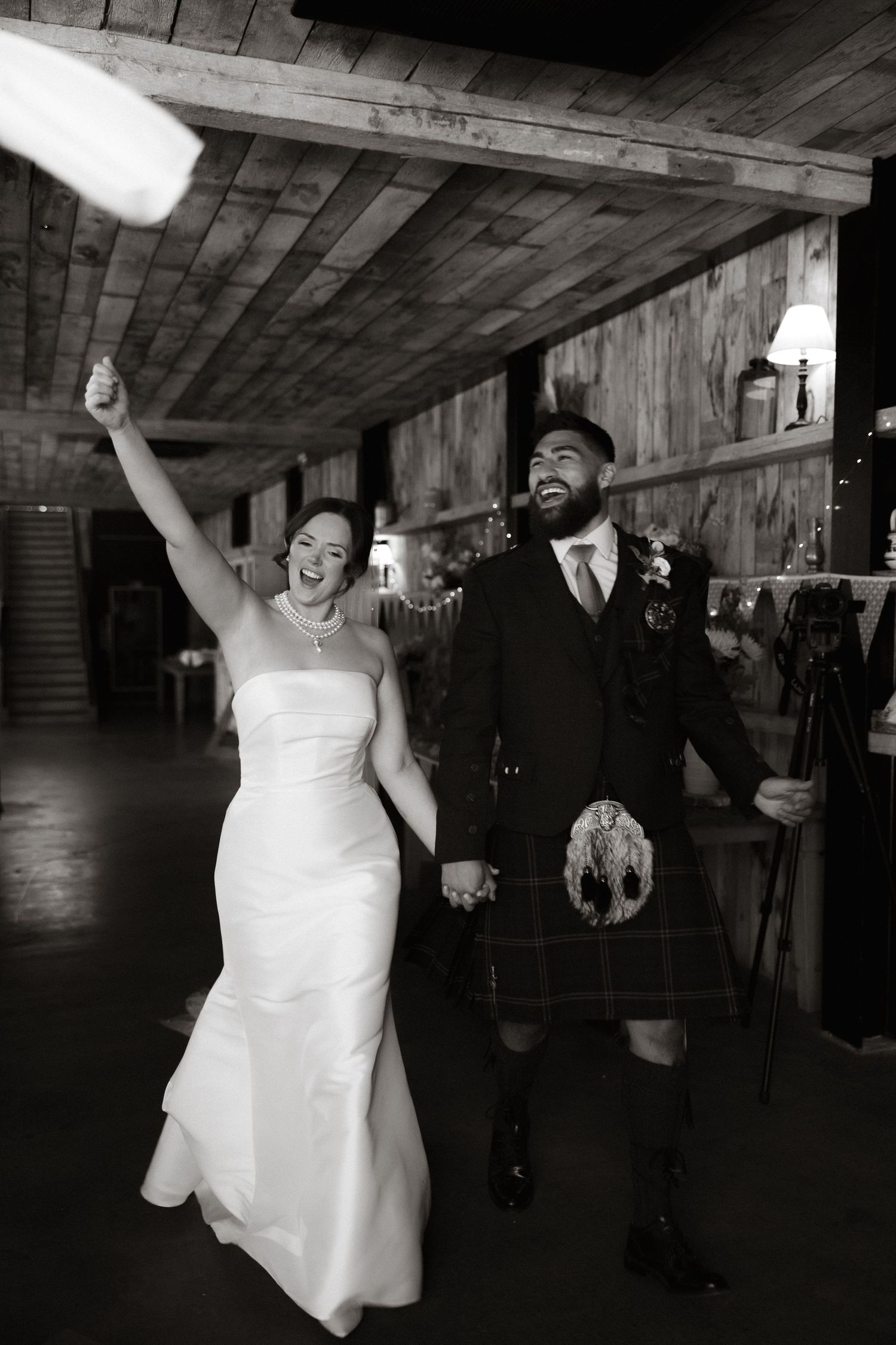 A wedding celebration in rustic wooden venue shows couple joyfully holding hands, with groom wearing traditional kilt.