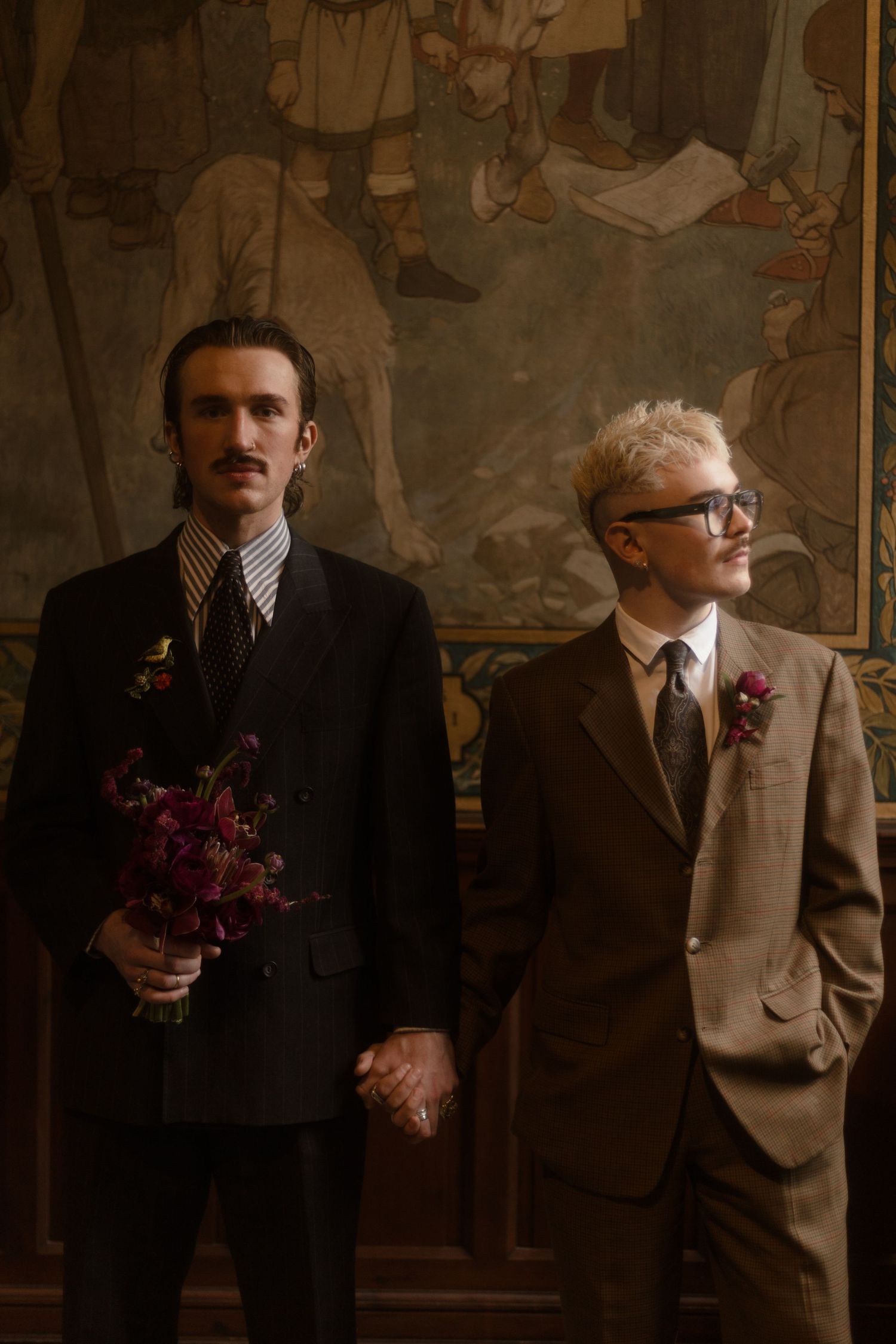 Two people in vintage suits stand together in a dimly lit room with dark wall art behind them.