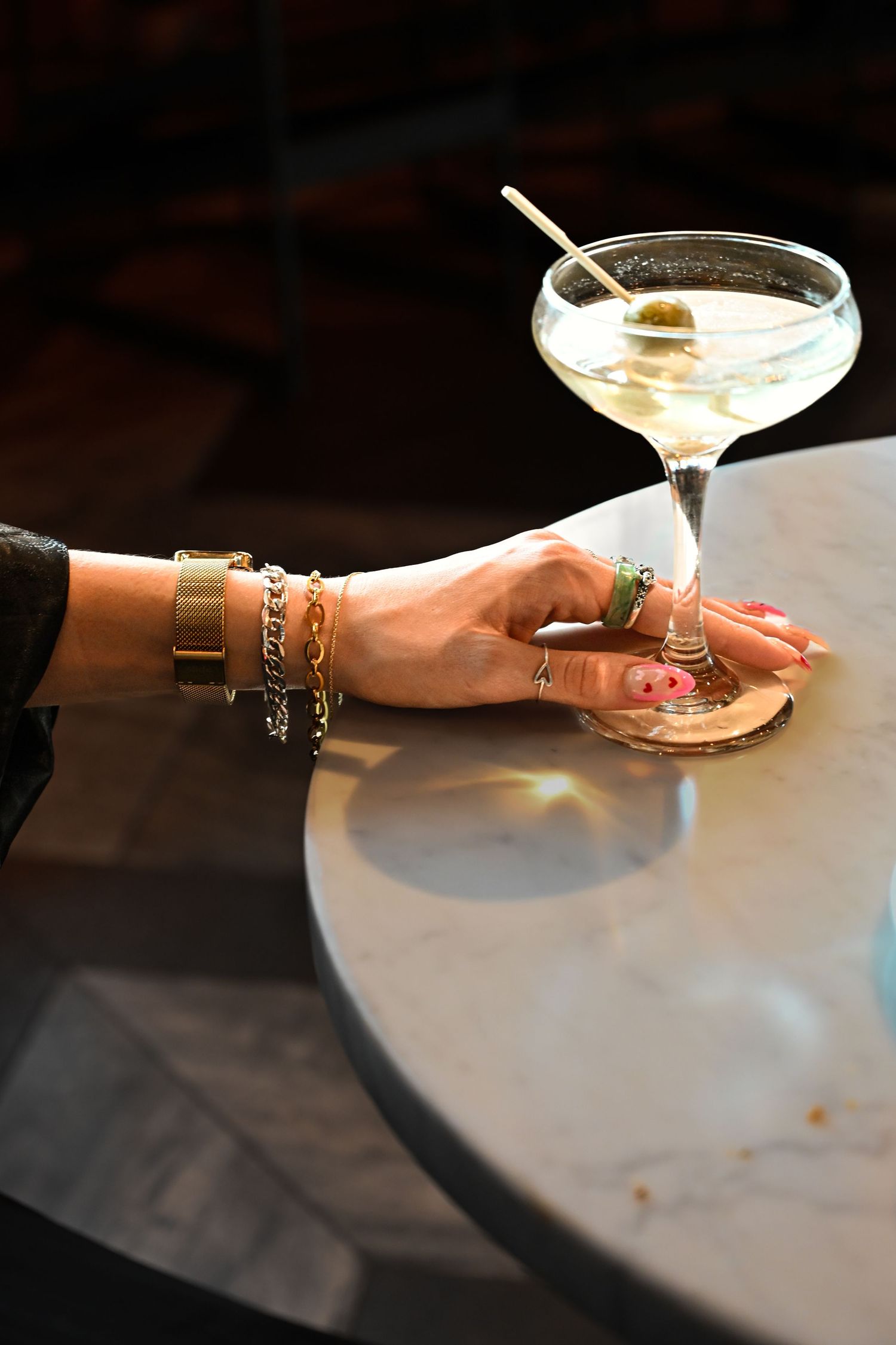 A manicured hand adorned with bracelets and rings reaches for a champagne coupe cocktail on a marble table.