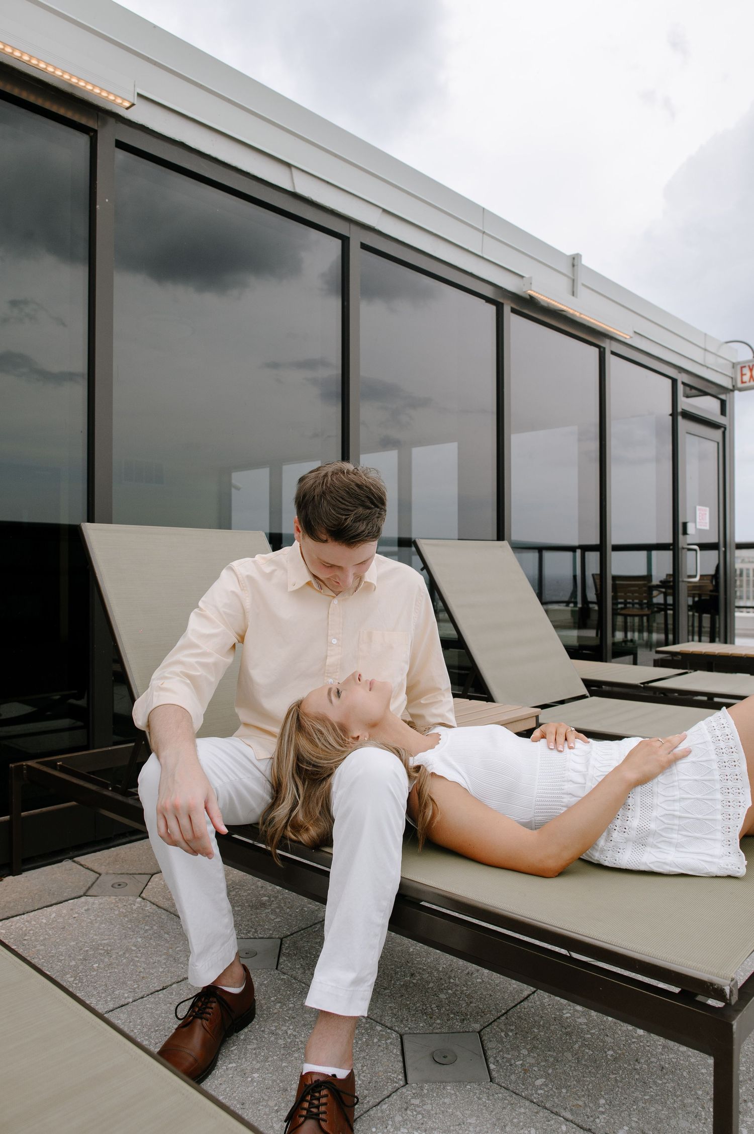 A person in white pants sits and reclines on a lounge chair on a modern terrace with glass windows.